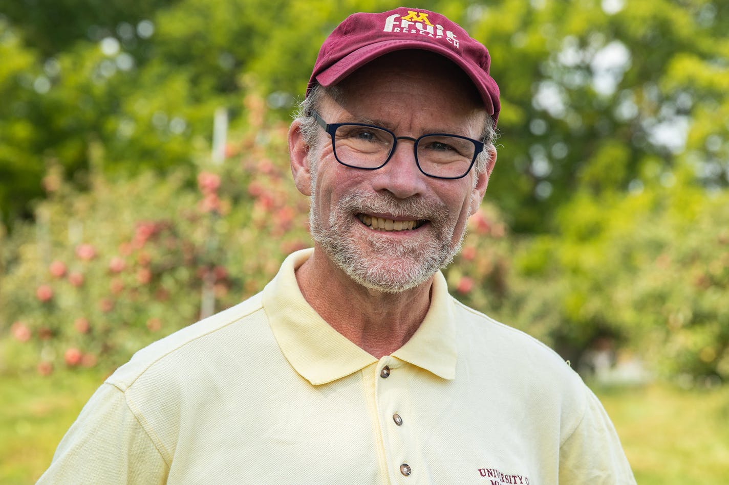 A middle-aged man wearing glasses and a short gray beard and a maroon hat smiles at the camera in front of an apple tree.