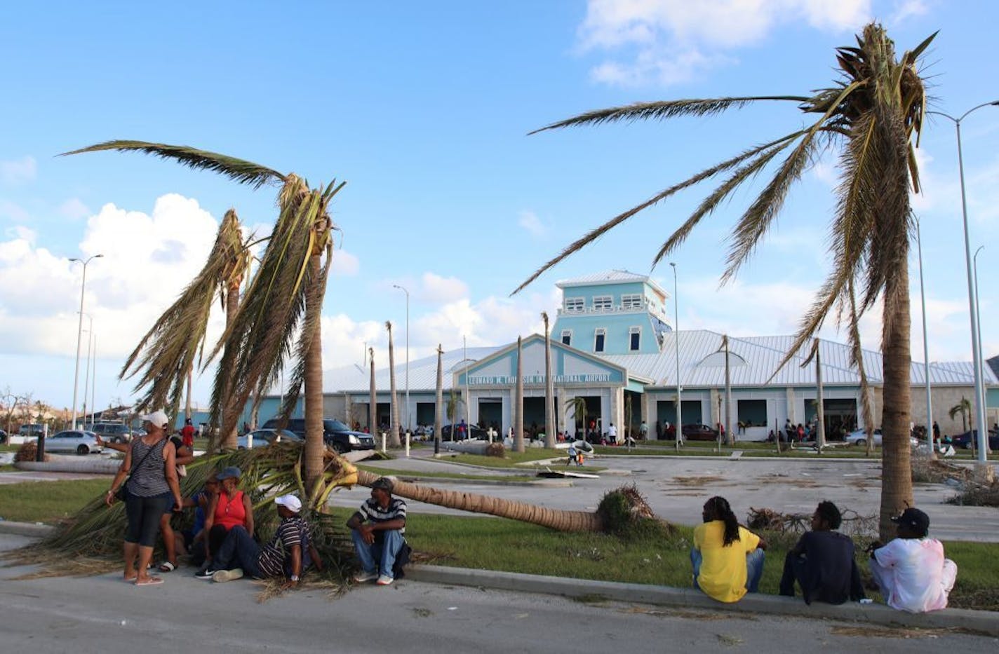 People sit under broken palm trees outside the Leonard M. Thompson International Airport after the passing of Hurricane Dorian in Marsh Harbour, Abaco Islands, Bahamas, Thursday, Sept. 5, 2019. Thousands of desperate people are seeking help in Dorian's aftermath.