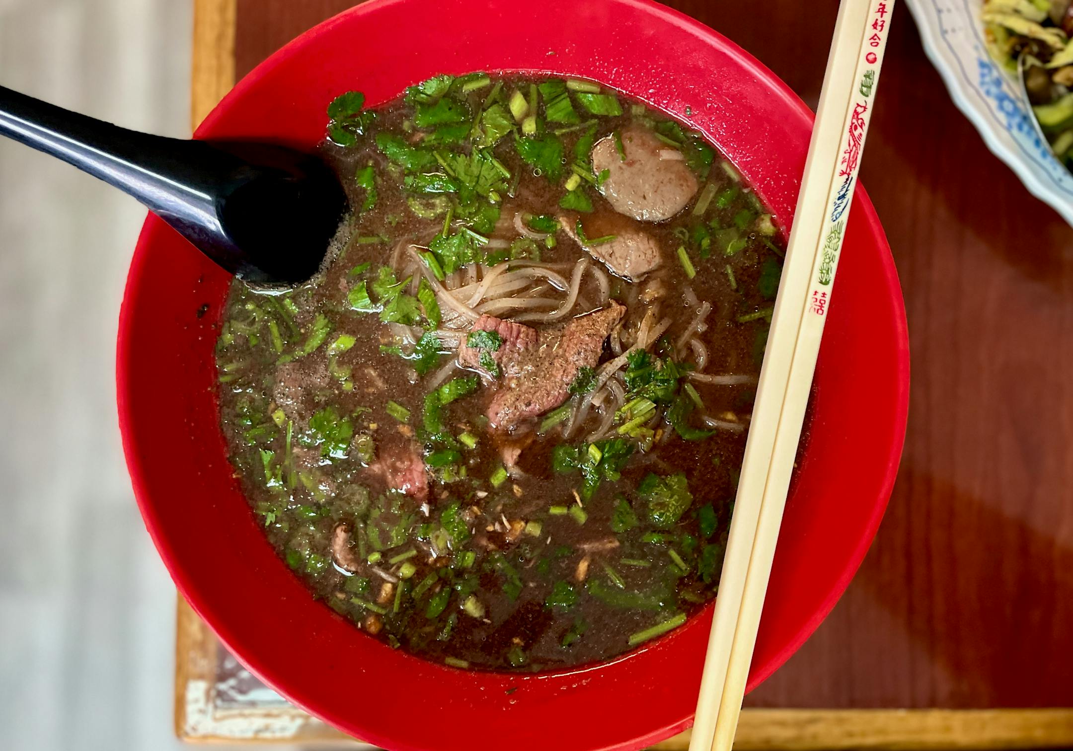 The large red bowl of deeply colored beef broth flecked with green herbs on a laminate table. On top of the bowl are balanced a spoon and chopsticks.