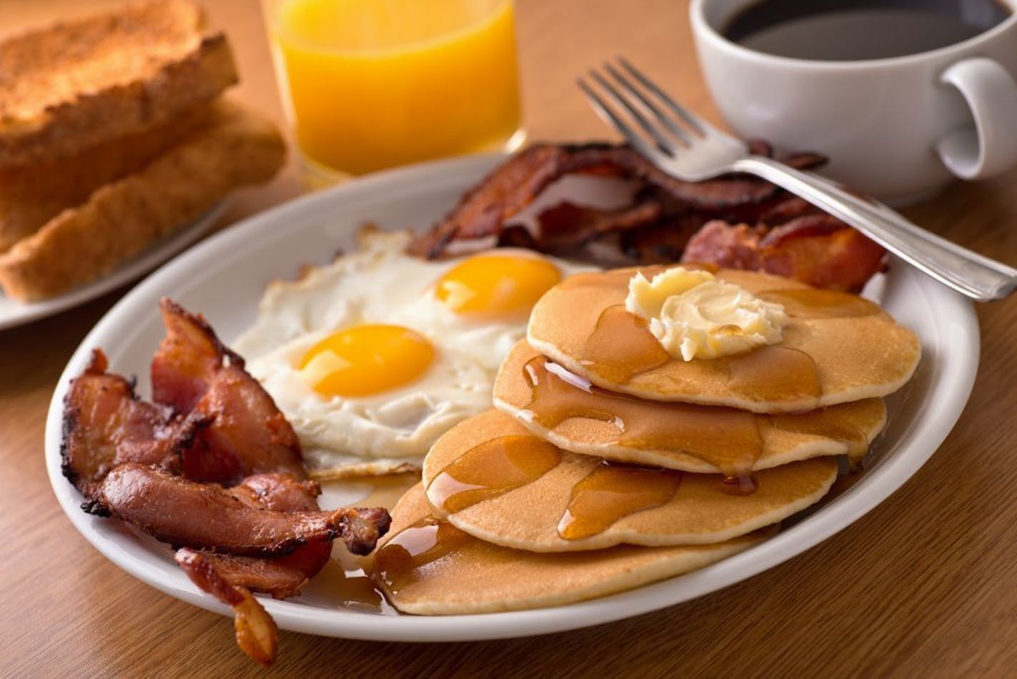 A delicious home style breakfast with crispy bacon, eggs, pancakes, toast, coffee and orange juice.