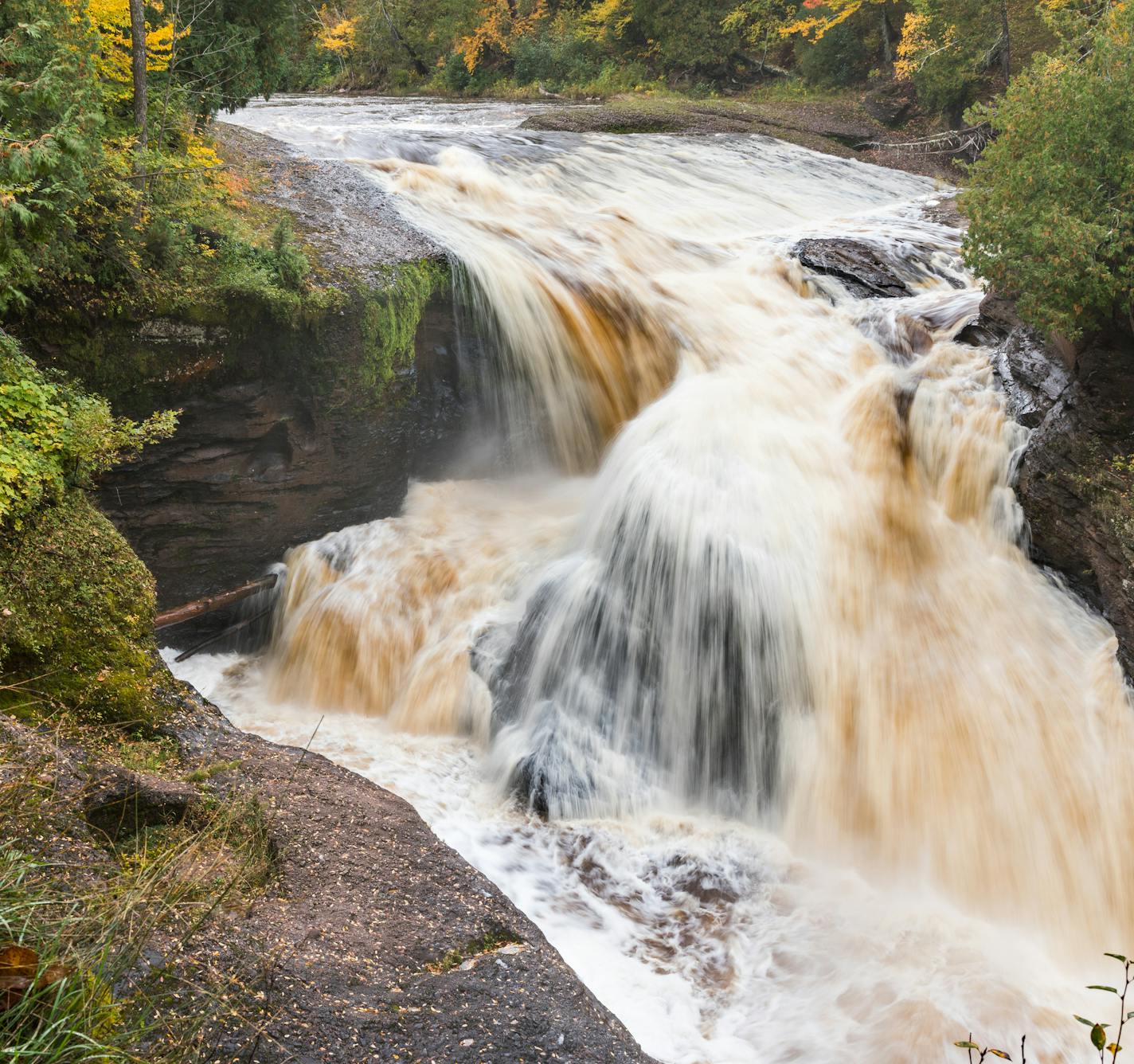 The scenic Black River rushes over Rainbow Falls in the Upper Peninsula of Michigan. The waterfall is surrounded by autumn colors.