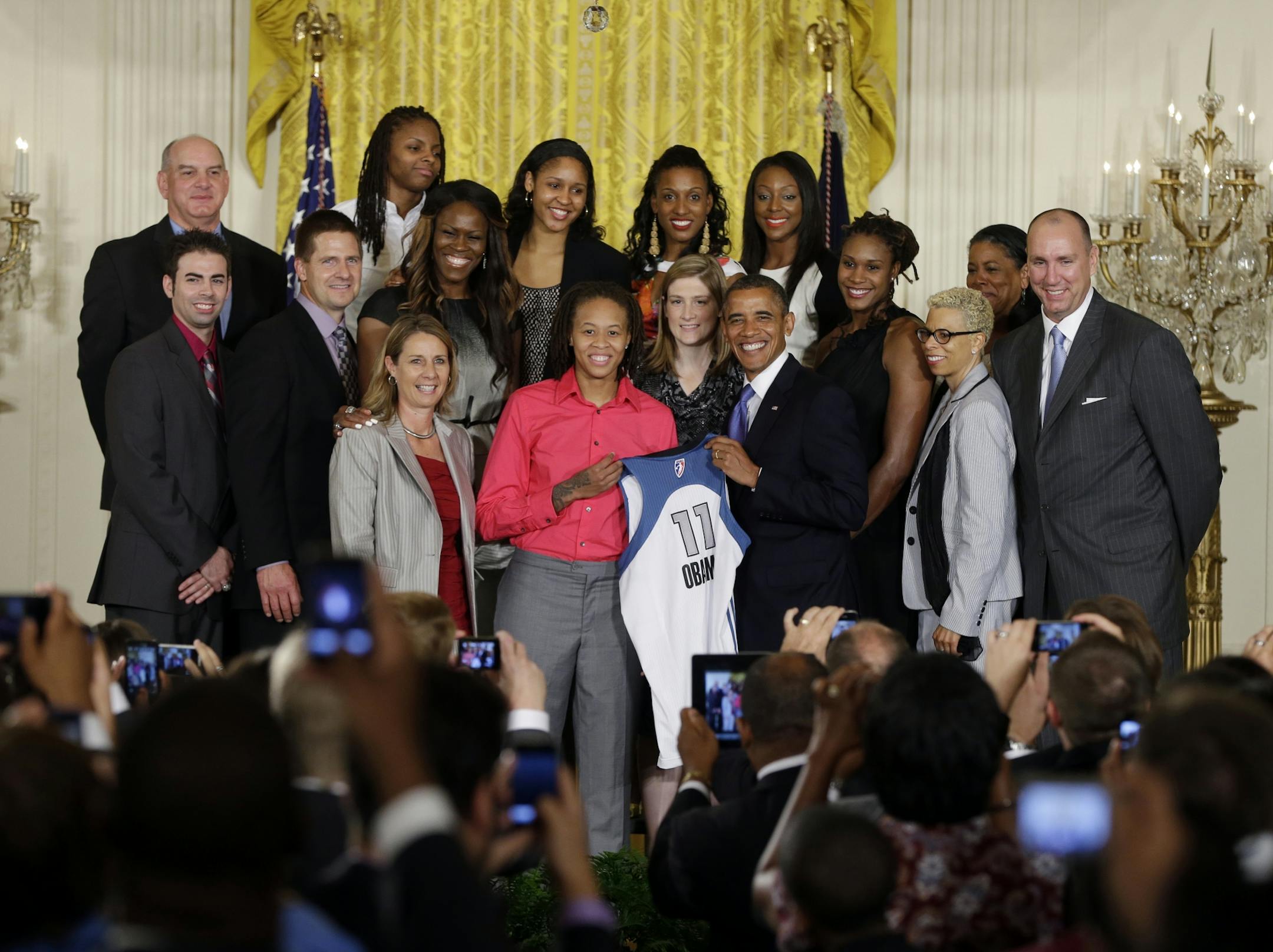 President Barack Obama, center right, holds up a Lynx team basketball jersey presented to him by Seimone Augustus, center left, as he honors the WNBA Champion Minnesota Lynx, Tuesday, Sept. 18, 2012, in a ceremony in the East Room of the White House in Washington.
