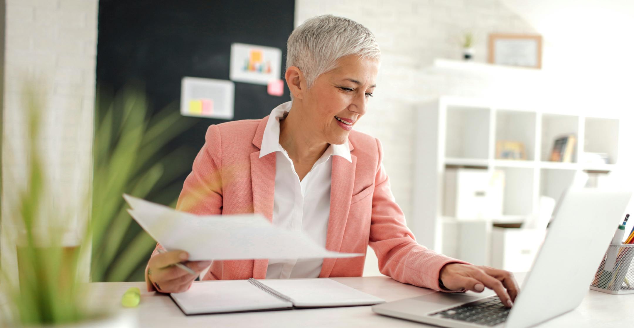 Mature businesswoman working in her new modern office. Sitting, examining documents and using laptop. She is focused on her work. Holding documents in her hand. istock