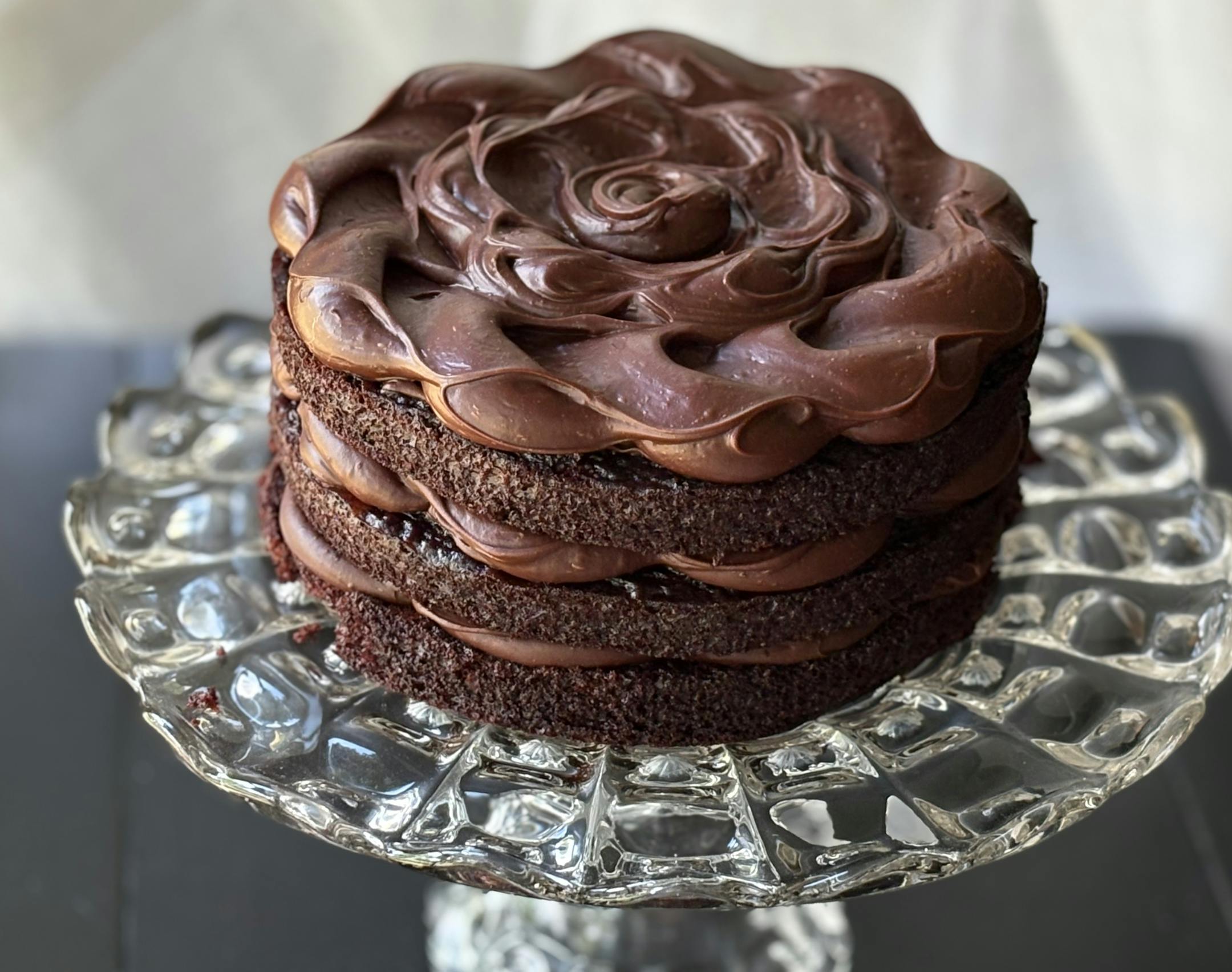 A three layer chocolate cake with chocolate frosting photographed on a clear glass cake stand.