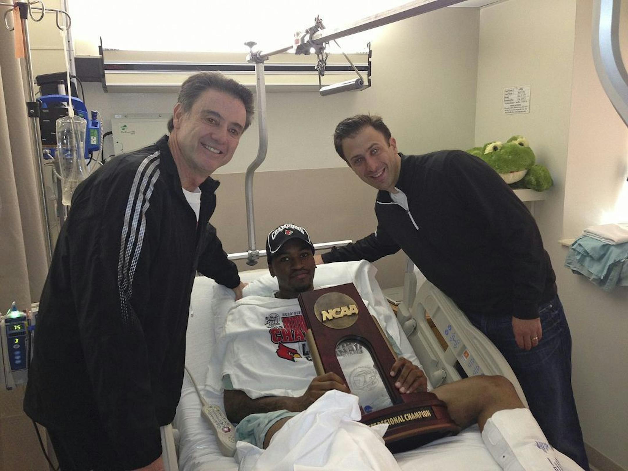 In this photo released by the University of Louisville, injured Louisville guard Kevin Ware lies in a hospital bed holding the NCAA Regional Championship trophy with coach Rick Pitino, left.