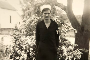 Black and white photo of Robert Kaping after he enlisted in the U.S. Navy, 1942, standing in a yard in front of a flowering bush.