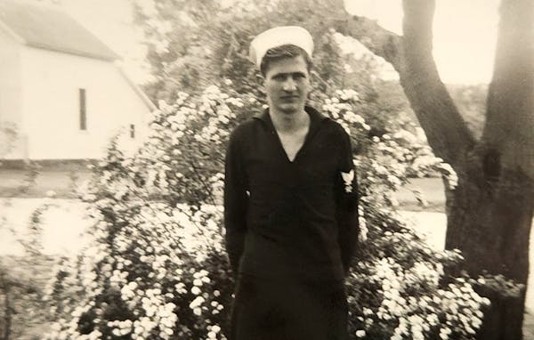 Black and white photo of Robert Kaping after he enlisted in the U.S. Navy, 1942, standing in a yard in front of a flowering bush.