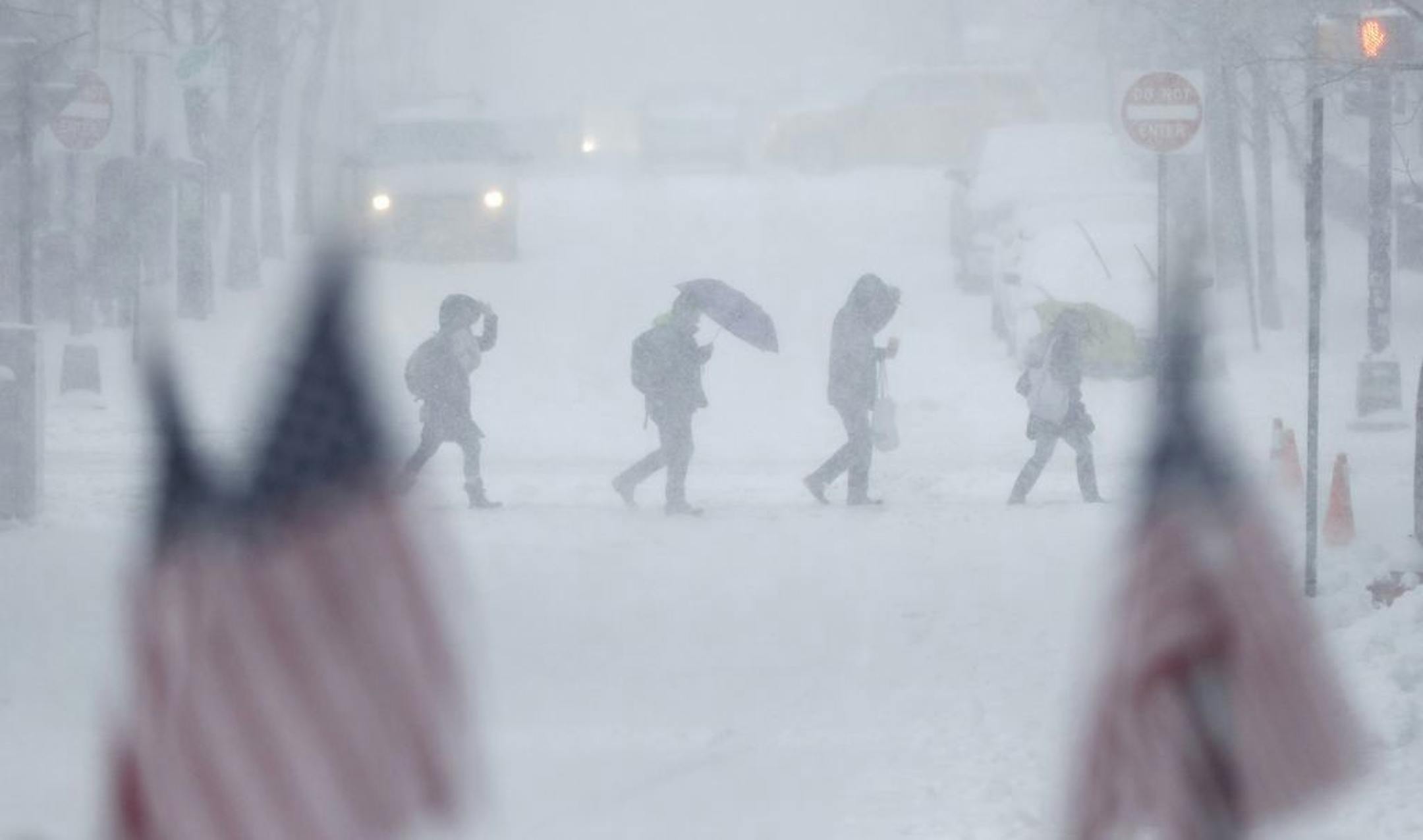 People walk across a snow-covered intersection behind American flags in Hoboken, N.J., Thursday, Feb. 9, 2017. A powerful, fast-moving storm swept through the northeastern U.S. Thursday, making for a slippery morning commute and leaving some residents bracing for blizzard conditions.