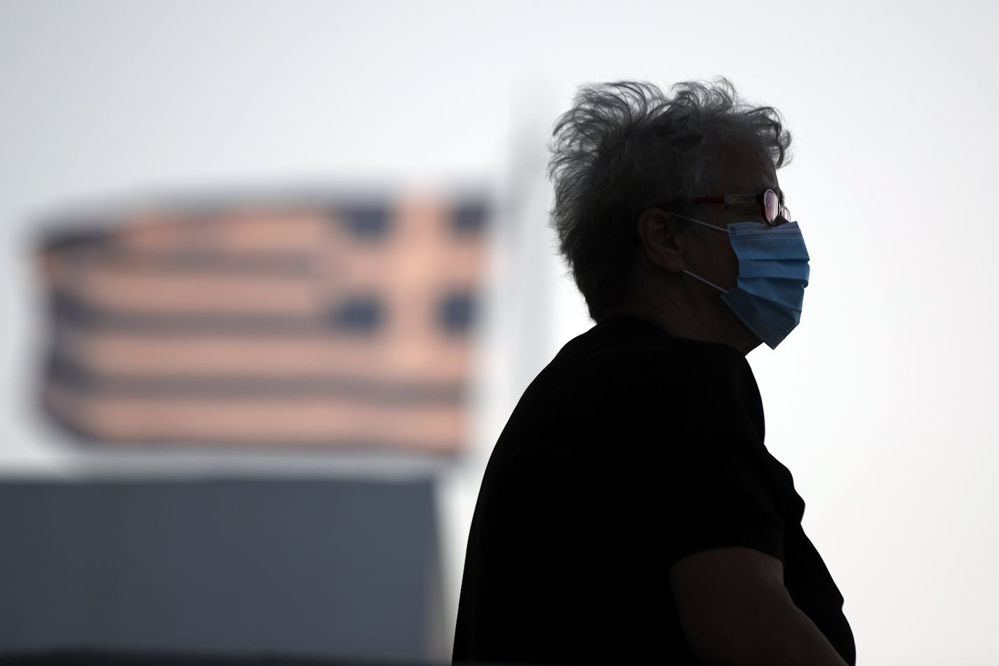 A elderly woman wearing a face mask sits outside a church as a Greek flag waves in the background, on the Aegean island of Tinos, Greece, on Thursday, Aug. 13, 2020. The country has suffered a spike in COVID-19 infections in recent days, largely attributed to lax protection measures among holiday pleasure-seekers. (AP Photo/Thanassis Stavrakis)