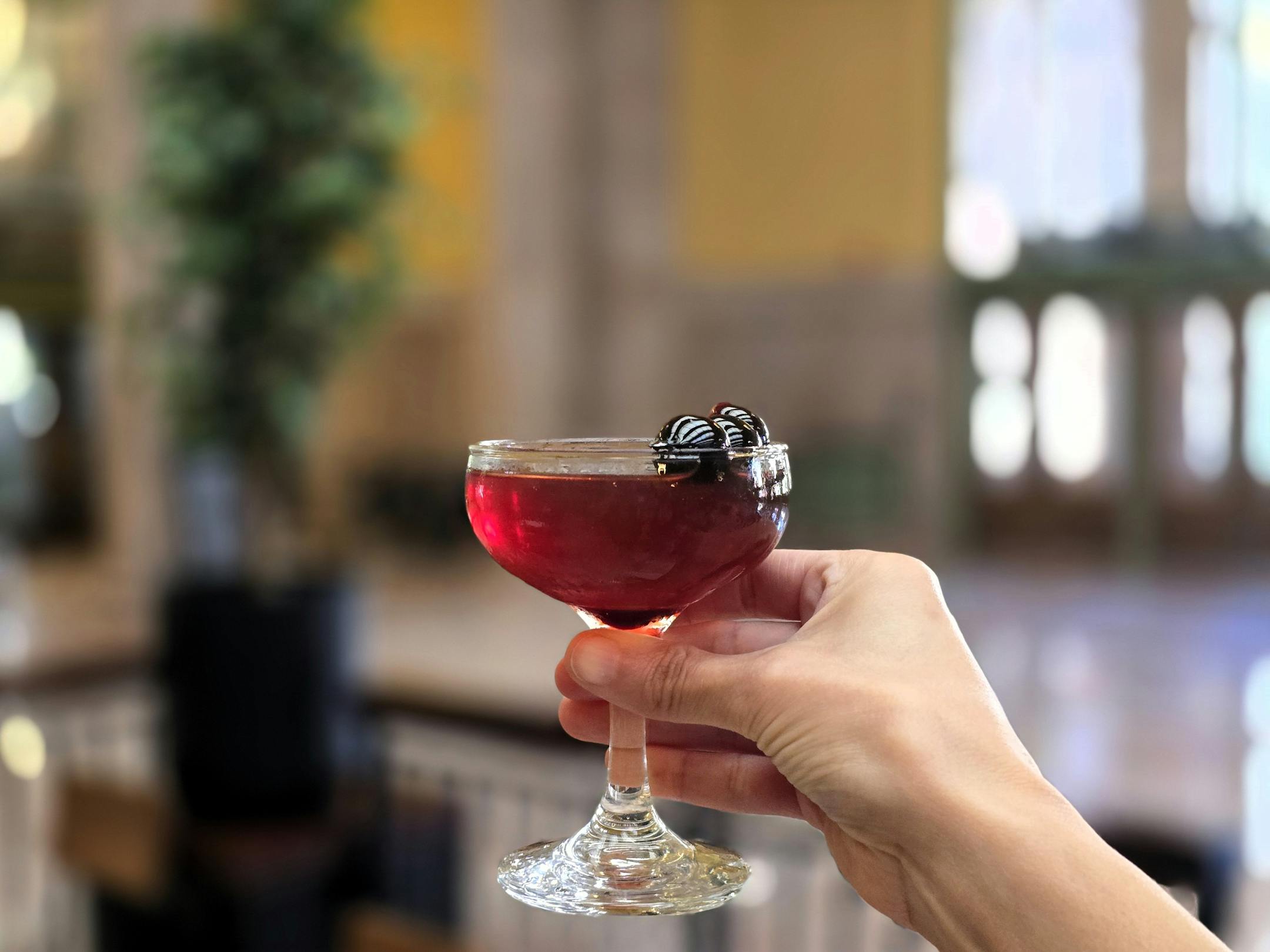 A hand holds up a reddish drink in a coupe glass with a skewer of cherries resting on top, in front of an open space with a yellow wall
