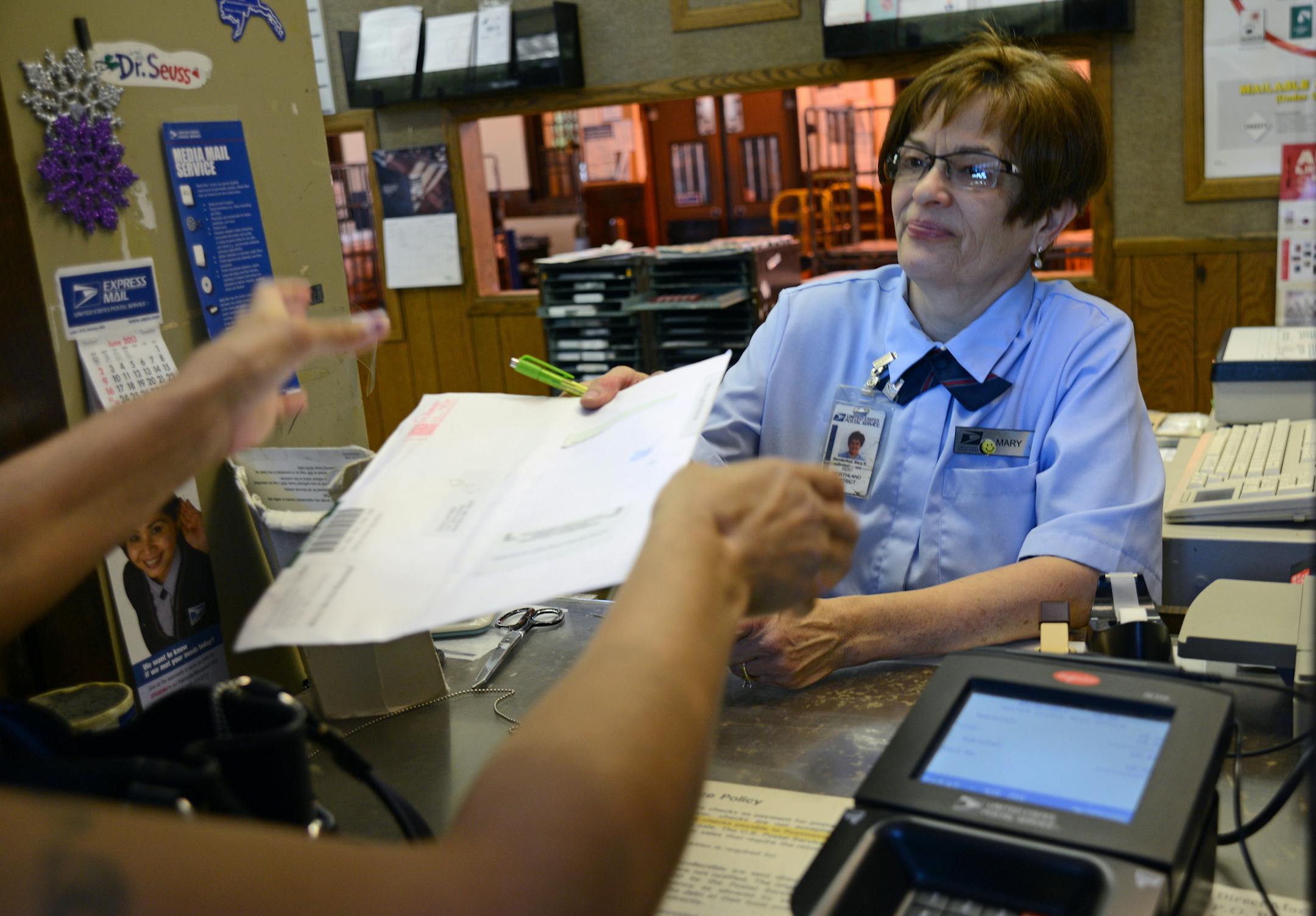 Mary Manderfeld, works behind the counter and places cut flowers in the lobby every day. The iris and lilacs grow behind the post office .The USPS has had the downtown Northfield post office for sale for more than two years, and the local Save our Post Office group, historic preservation groups, and the community are concerned because the post office has not started the regulatory process (Section 106) and studying impact of giving up ownership of a historic and federal site, which requires not