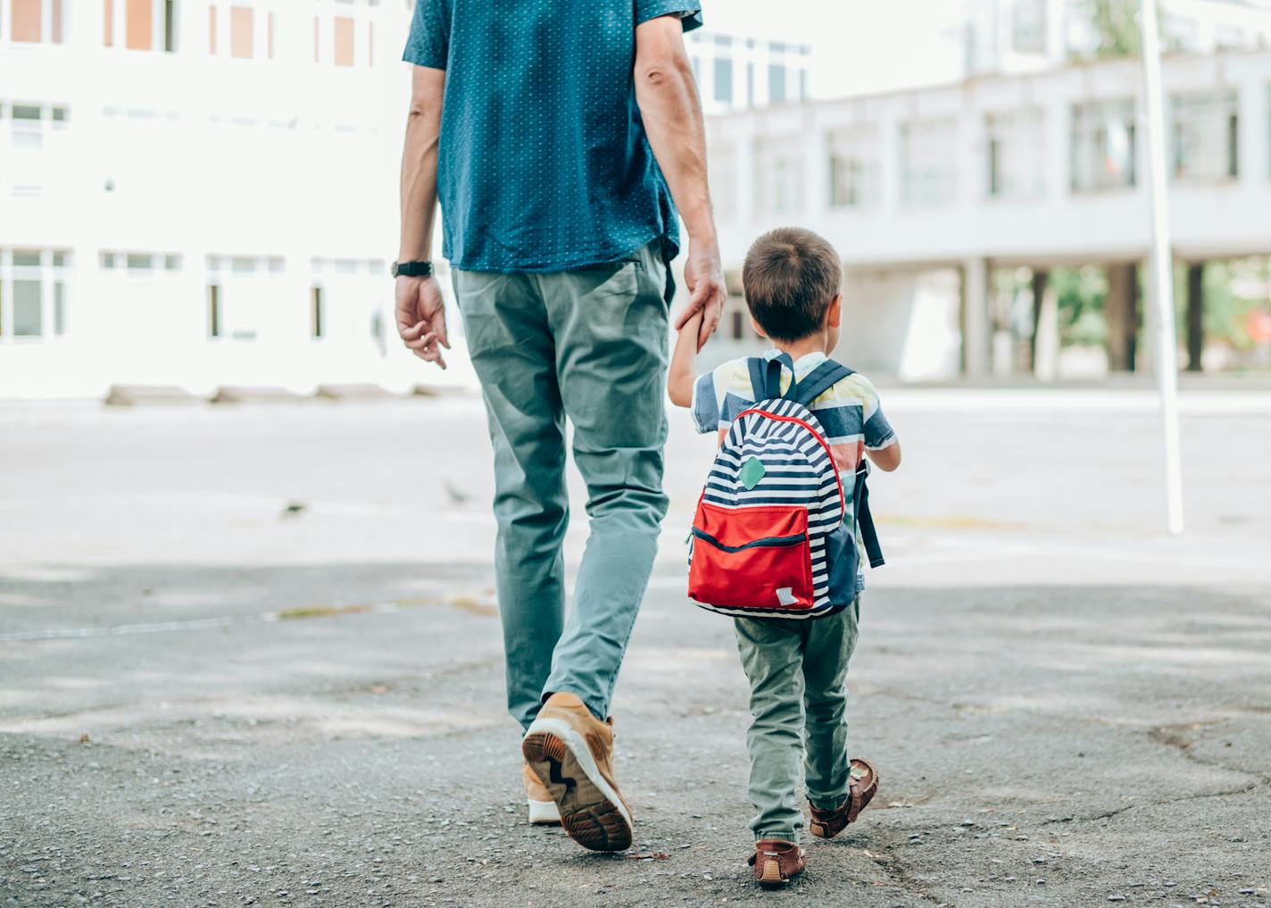 Rear view of father who leads a little boy hand in hand to the school. Father and son with backpack walking in schoolyard.