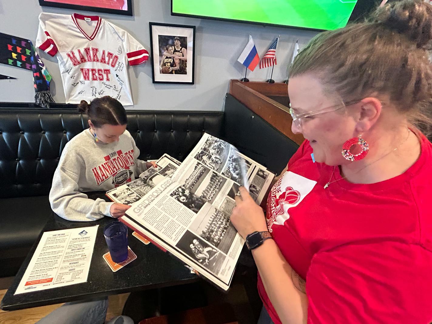 Two women page through black-and-white photos of a yearbook, smiling, at a restaurant booth.