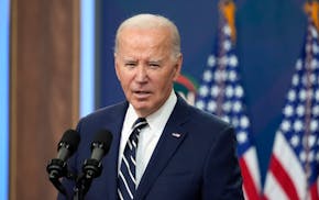 President Joe Biden wearing navy suit, white shirt and blue and white striped tie speaking into double microphone with U.S. flags in background.