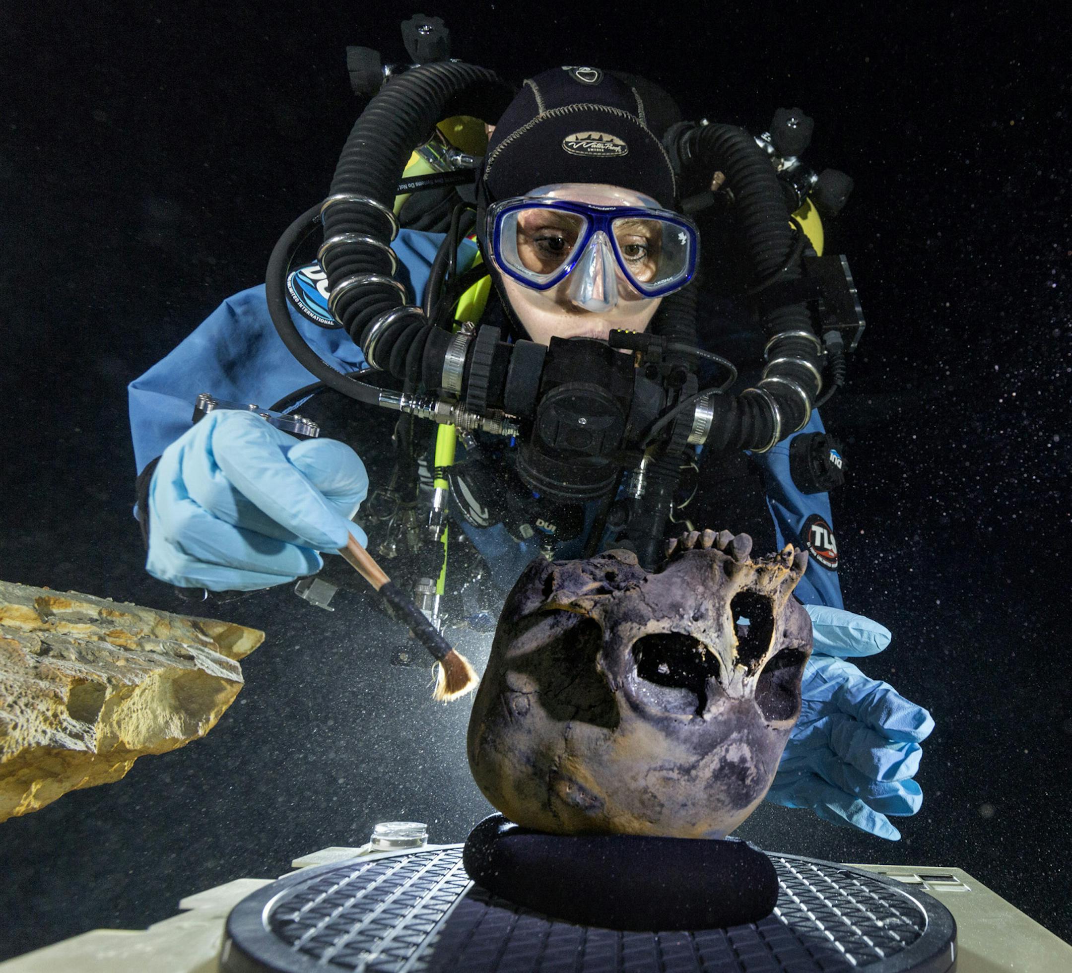 In this June 2013 photo provided by National Geographic, diver Susan Bird, working at the bottom of Hoyo Negro, a large dome-shaped underwater cave in Mexico's Yucatan Peninsula, brushes a human skull found at the site while her team members take detailed photographs. Thousands of years ago, a teenage girl fell into this deep hole and died. Now, her skeleton and her DNA are helping scientists study the origins of the first Americans. An analysis of her remains was released Thursday, May 15, 2014