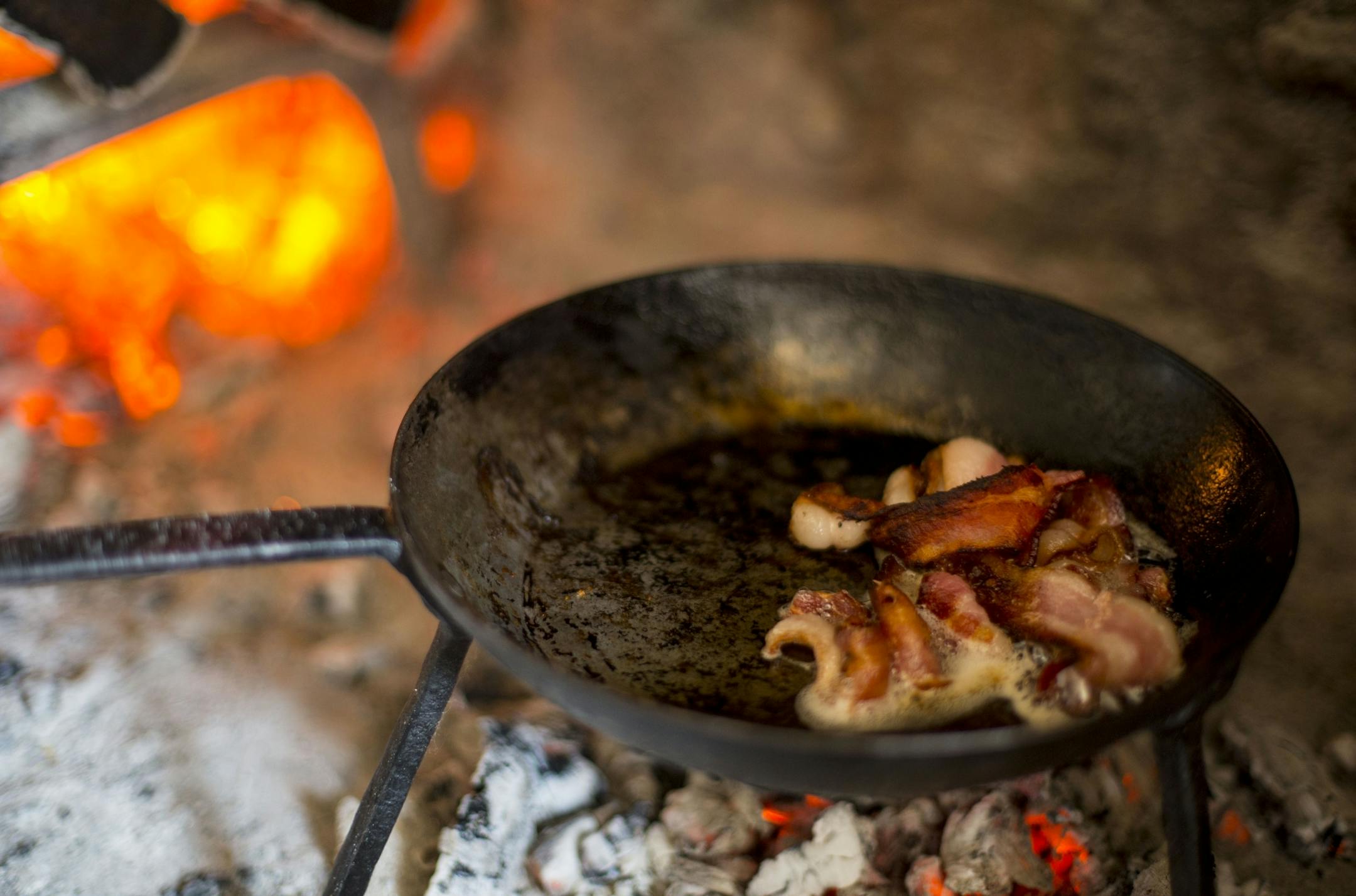 Bacon cooks on a small pan over a small fire of coals.