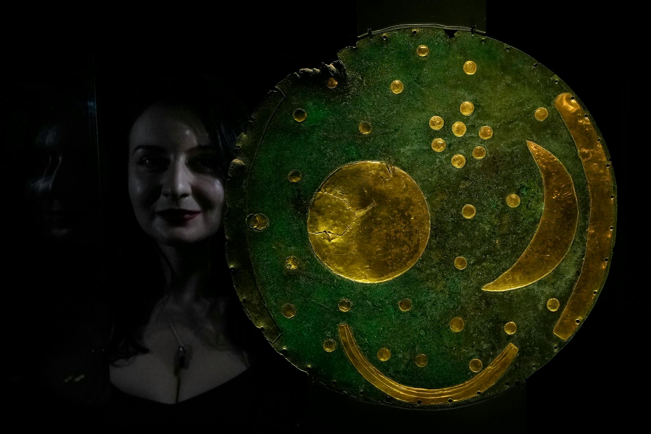 A member of staff poses next to the 'Nebra Sky Disc' which dates from around 1600 BCE, and is the oldest surviving representation of the cosmos, on display at the The World of Stonehenge' exhibition at the British Museum in London, Monday, Feb. 14, 2022. The exhibition which displays objects and artifacts from the era of Stonehenge opens 17, Feb. and runs until 17, July 2022. The Nebra Disc was found in Nebra in Saxony-Anhalt in east Germany, in 1999. (AP Photo/Alastair Grant)