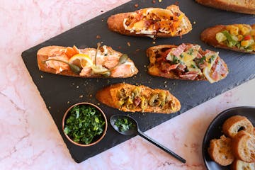 A black slate board with toasted bread with various toppings, making bruschetta for dinner.