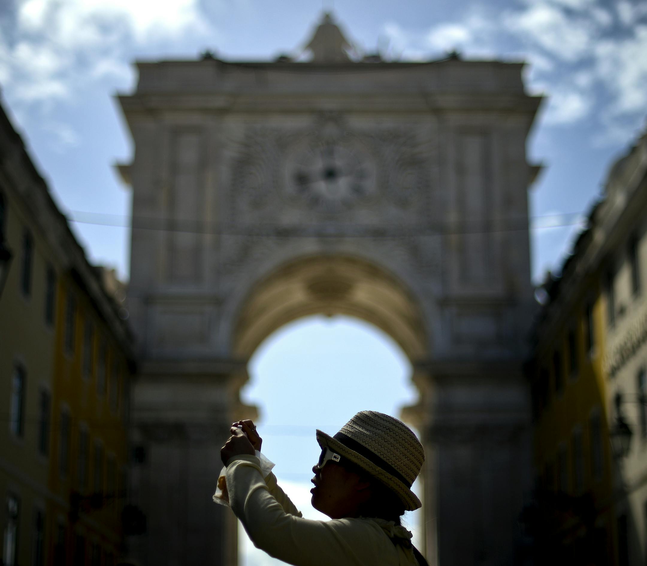 A tourist takes a picture under the Augusta Street Arch in sunny, warm Lisbon, Portugal. But it’s not home.