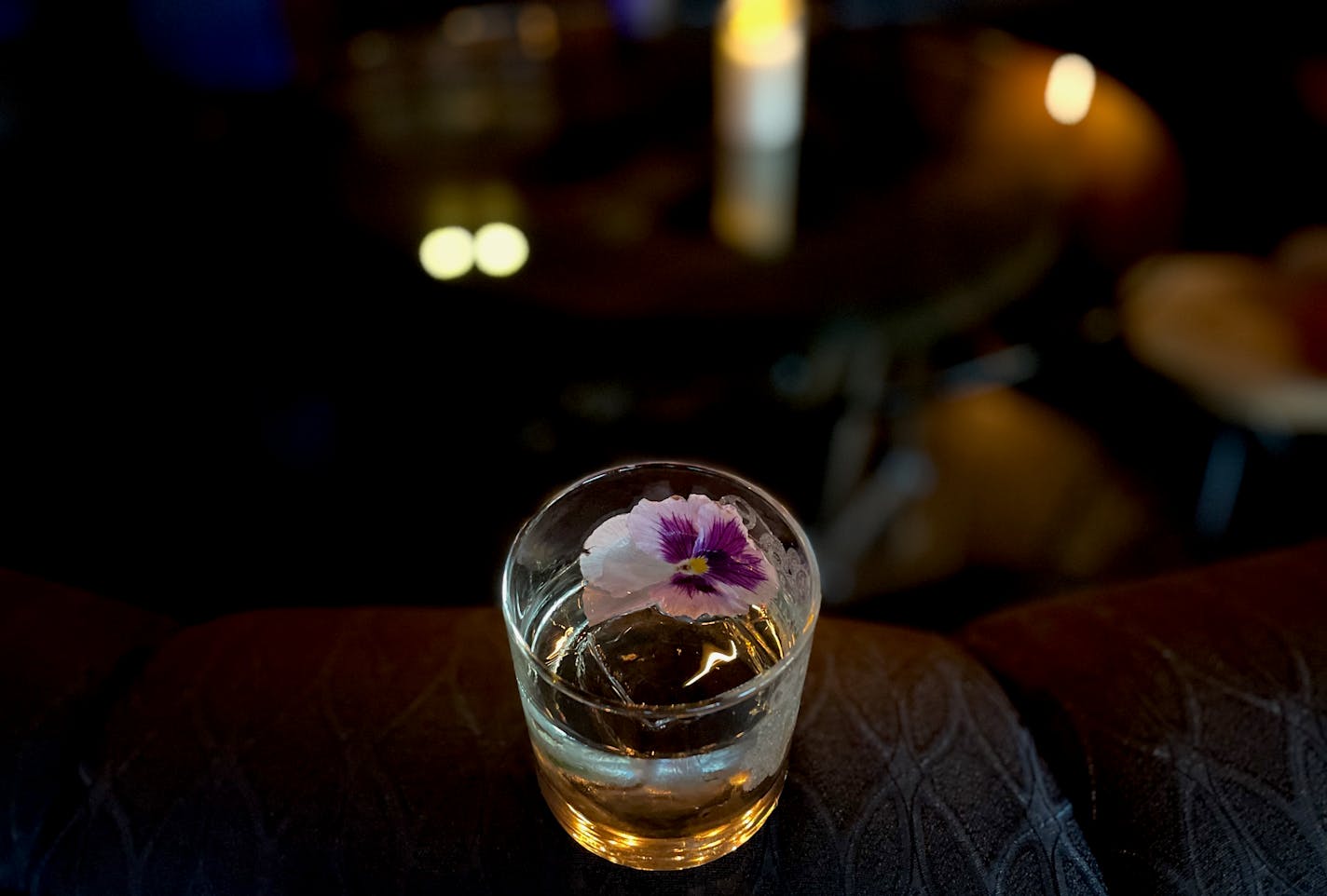 A clear cocktail balances on the back of a black leather couch, garnished with a white and purple pansy. In the background candles flicker.