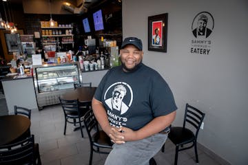 A Black man wearing a black t-shirt and gray jeans sits in front of a deli counter