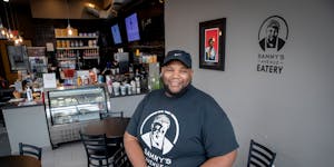 A Black man wearing a black t-shirt and gray jeans sits in front of a deli counter