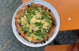 A charred crust pizza on a picnic table patio is topped with fresh salad greens and fried crispy cheese bits.