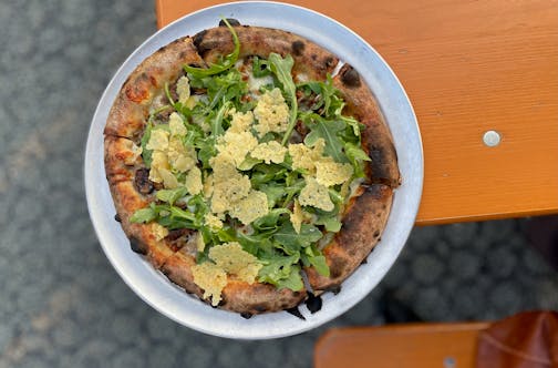 A charred crust pizza on a picnic table patio is topped with fresh salad greens and fried crispy cheese bits.