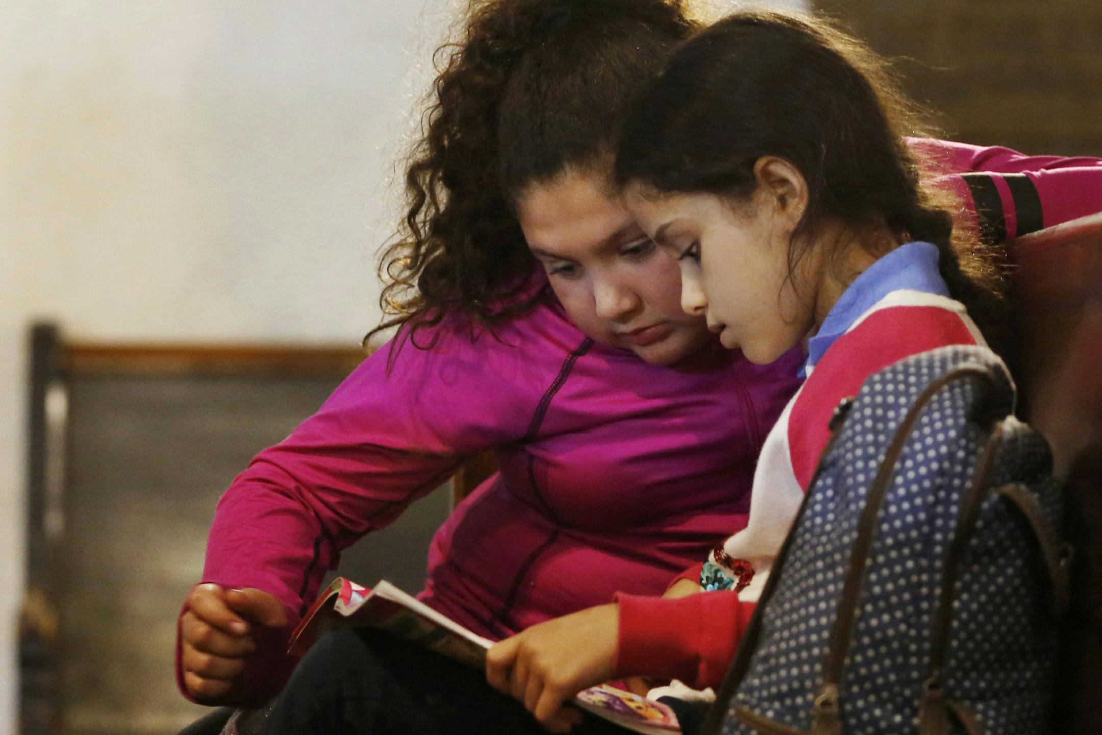 2 girls reading a book; one girl is wearing a fuchsia shirt and the other a red and white sweater over a blue shirt, with a backpack next to her.