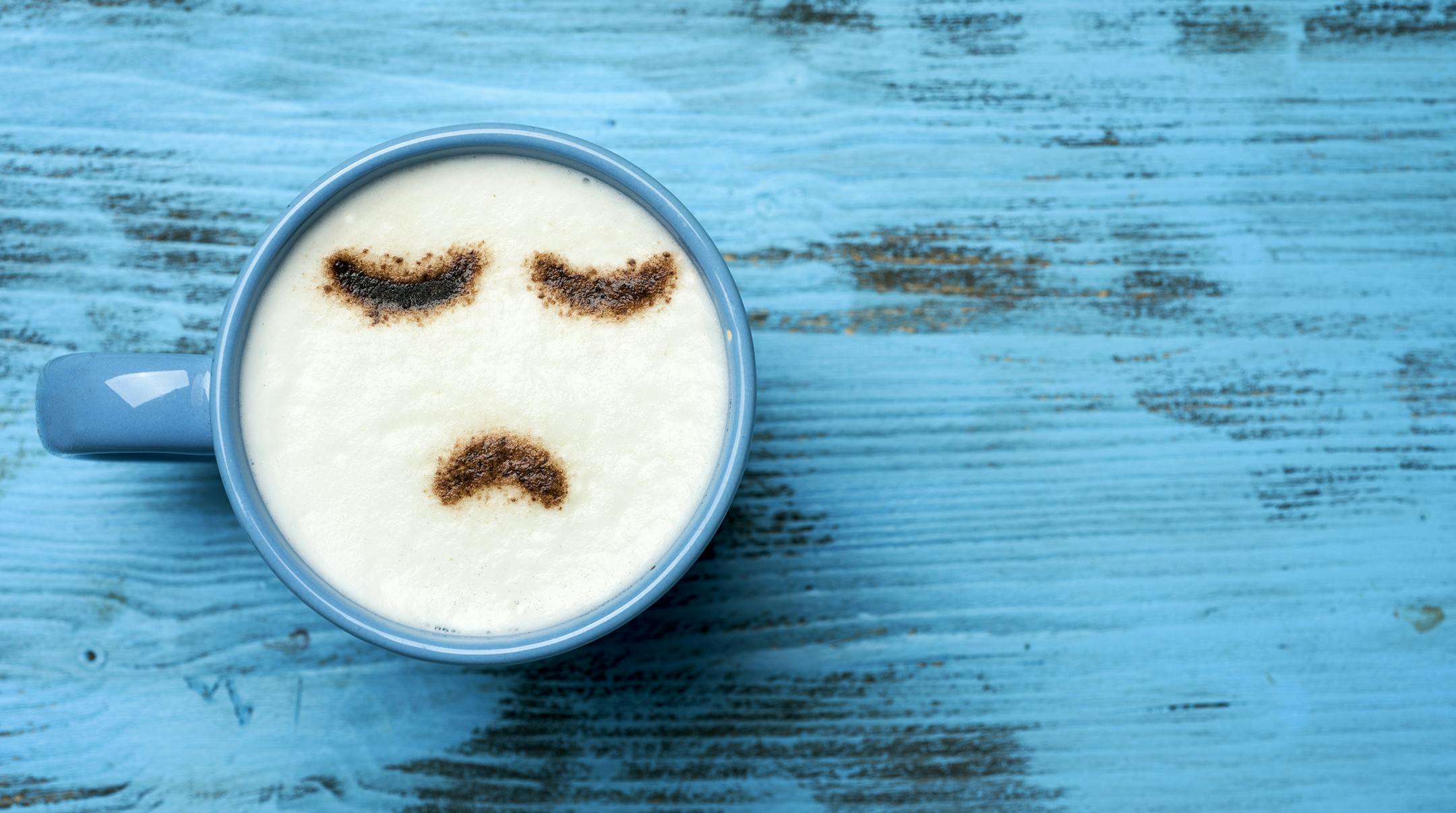 high-angle shot of a blue cup of cappuccino with a sad face drawn with cocoa powder on its milk foam, on a blue rustic table with a blank space on the right