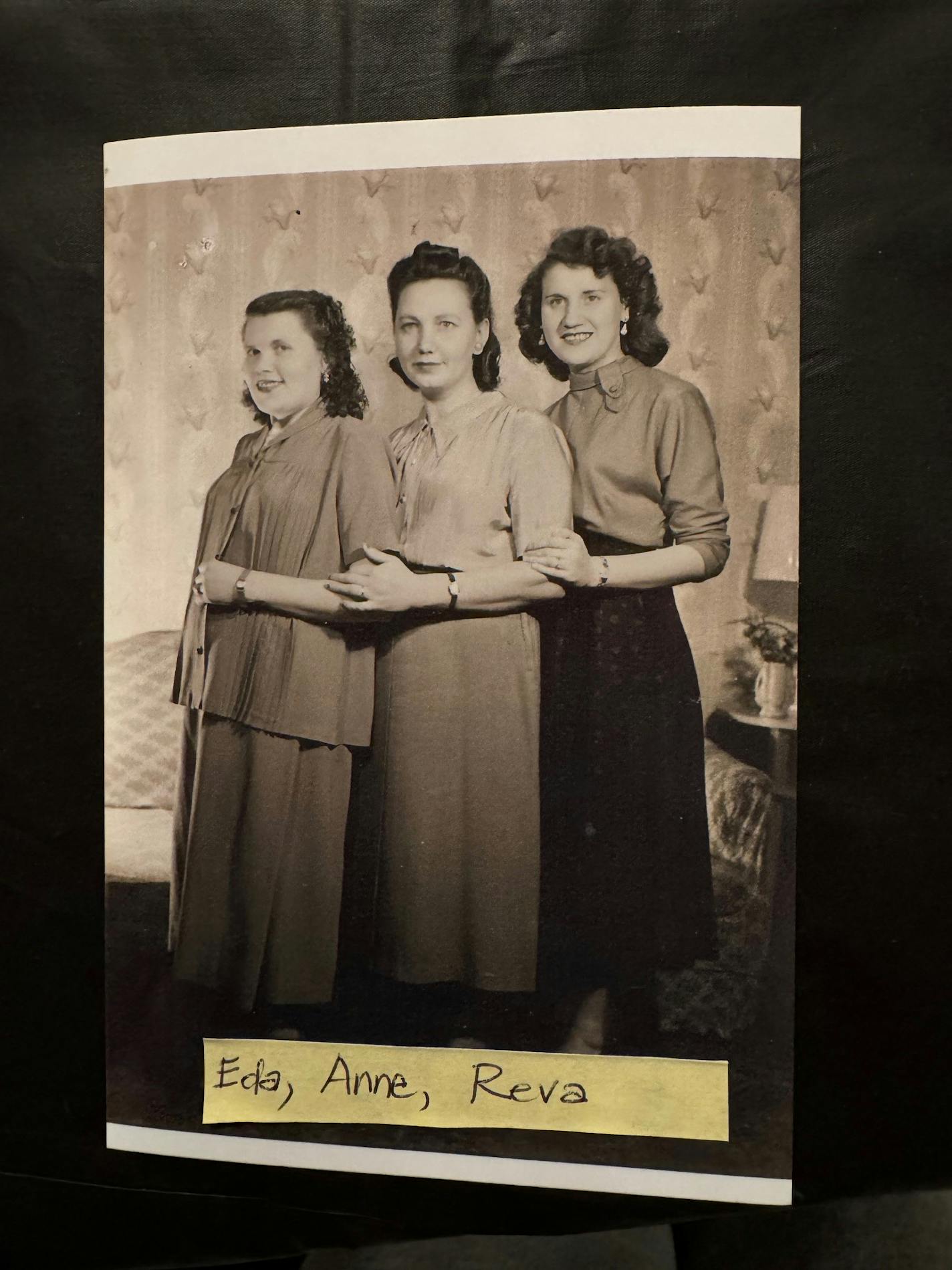 Three women wearing likely 1950s attire stand together with hands on the others' elbows.