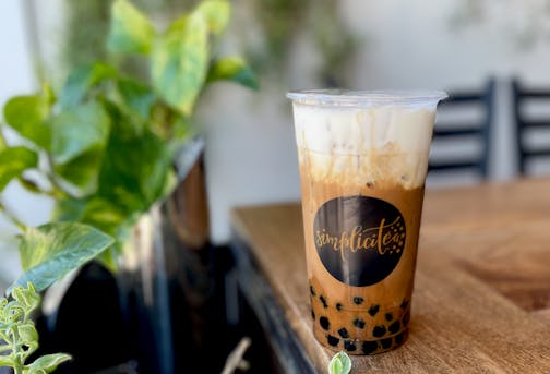 The separated layers of boba, coffee, foam are layered in a clear cup on a table surrounded by green plants.