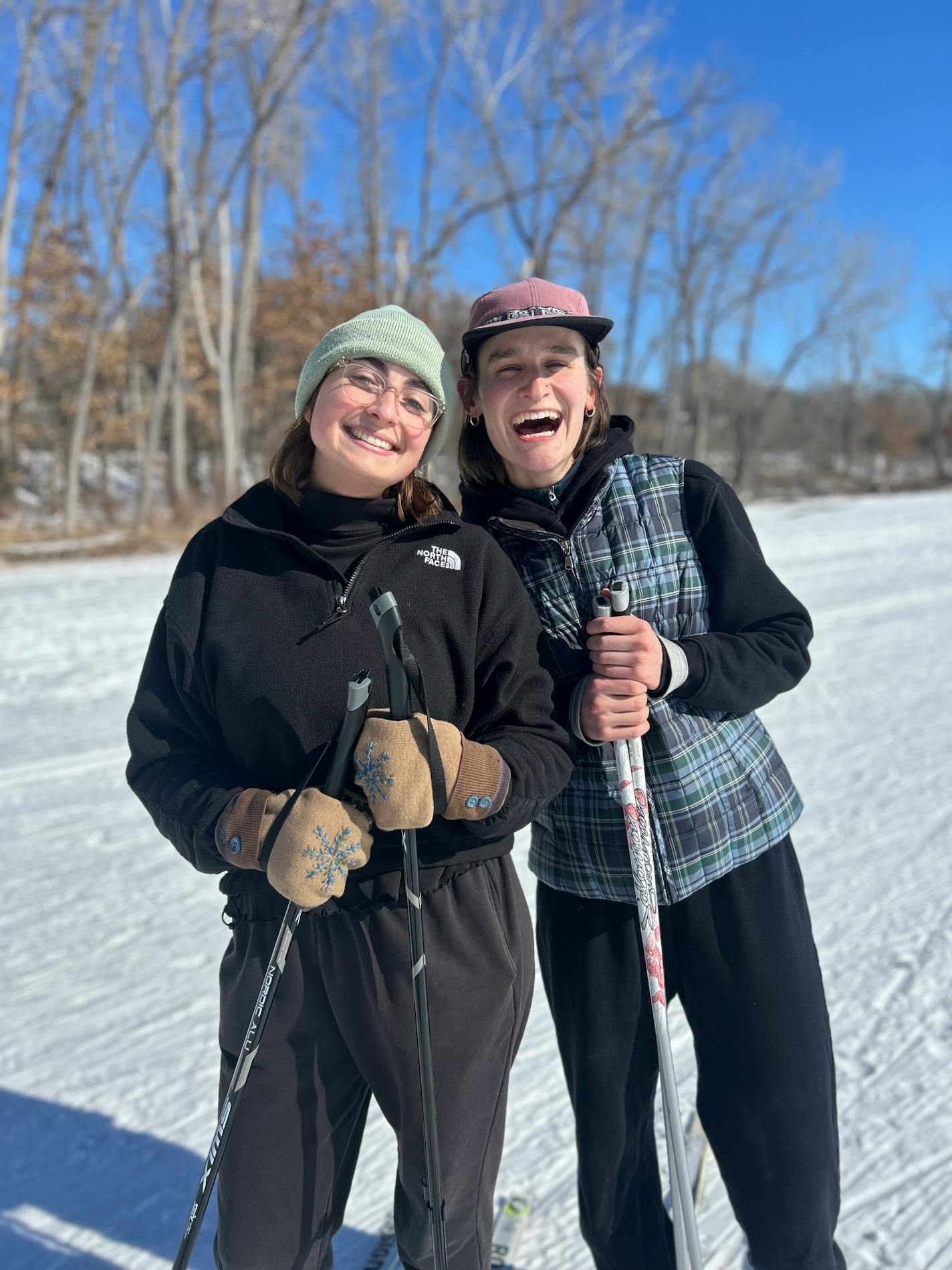 Two women in their 20s holding ski poles smile while standing on a frozen, snow-covered lake with bare trees in the background.