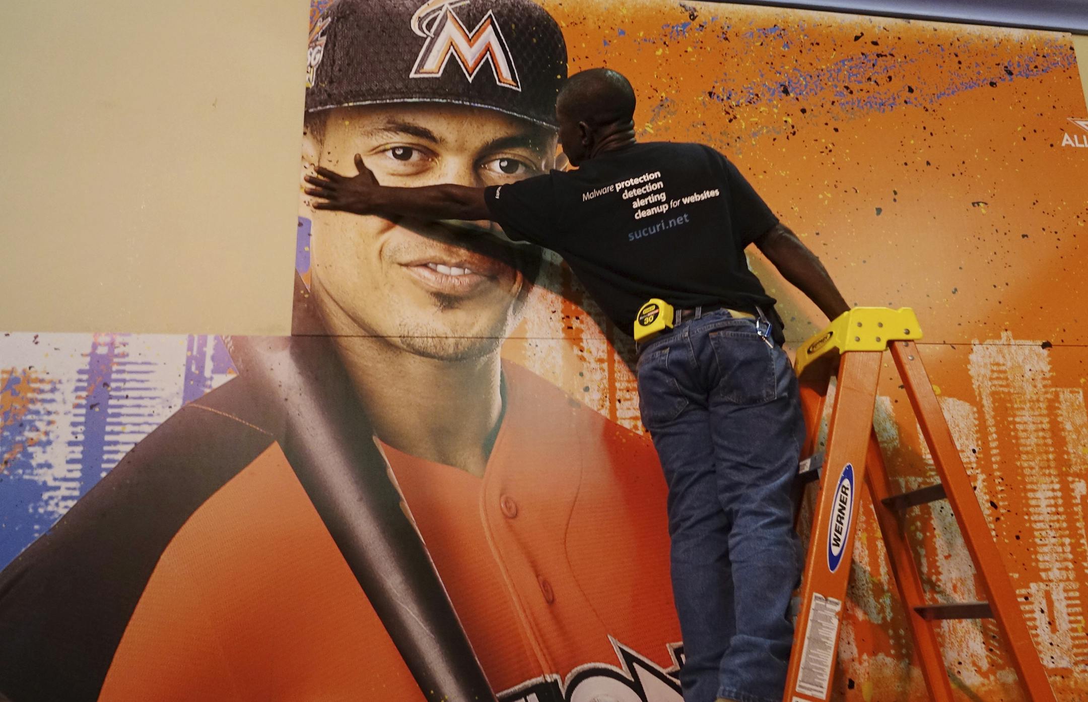 Francois Diesiul hangs a poster bearing the visage of Miami Marlins star Giancarlo Stanton, Thursday, July 6, 2017, at the Miami Beach Convention Center in Miami Beach, Fla., where baseball's All Star Game FanFest will open Friday morning. (Joe Cavaretta/South Florida Sun-Sentinel via AP)
