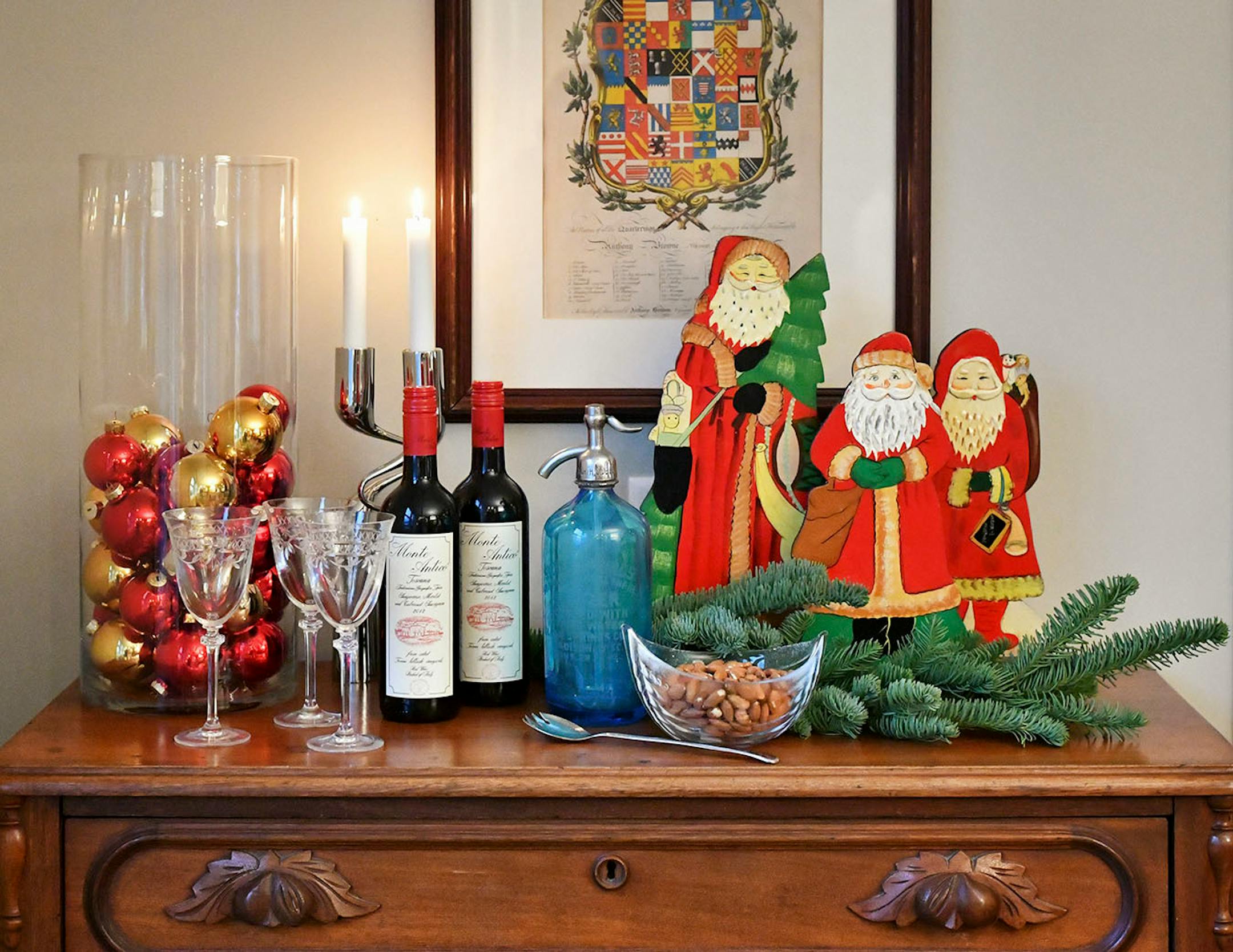 Euro Beverage Bar: Three colorful painted vintage Santas from Germany are the centerpiece. For the rest of the display, choose items with a variety of finishes and textures, such as a vintage glass seltzer bottle, shiny chrome Danish candleholder and cut crystal wine glasses and nut bowl. Partially fill a tall hurricane vase with red and gold glass balls, repeating the colors in the vintage Santas. To anchor the arrangement, Jay Nuhring hung artwork of a European coat of arms above the buffet.