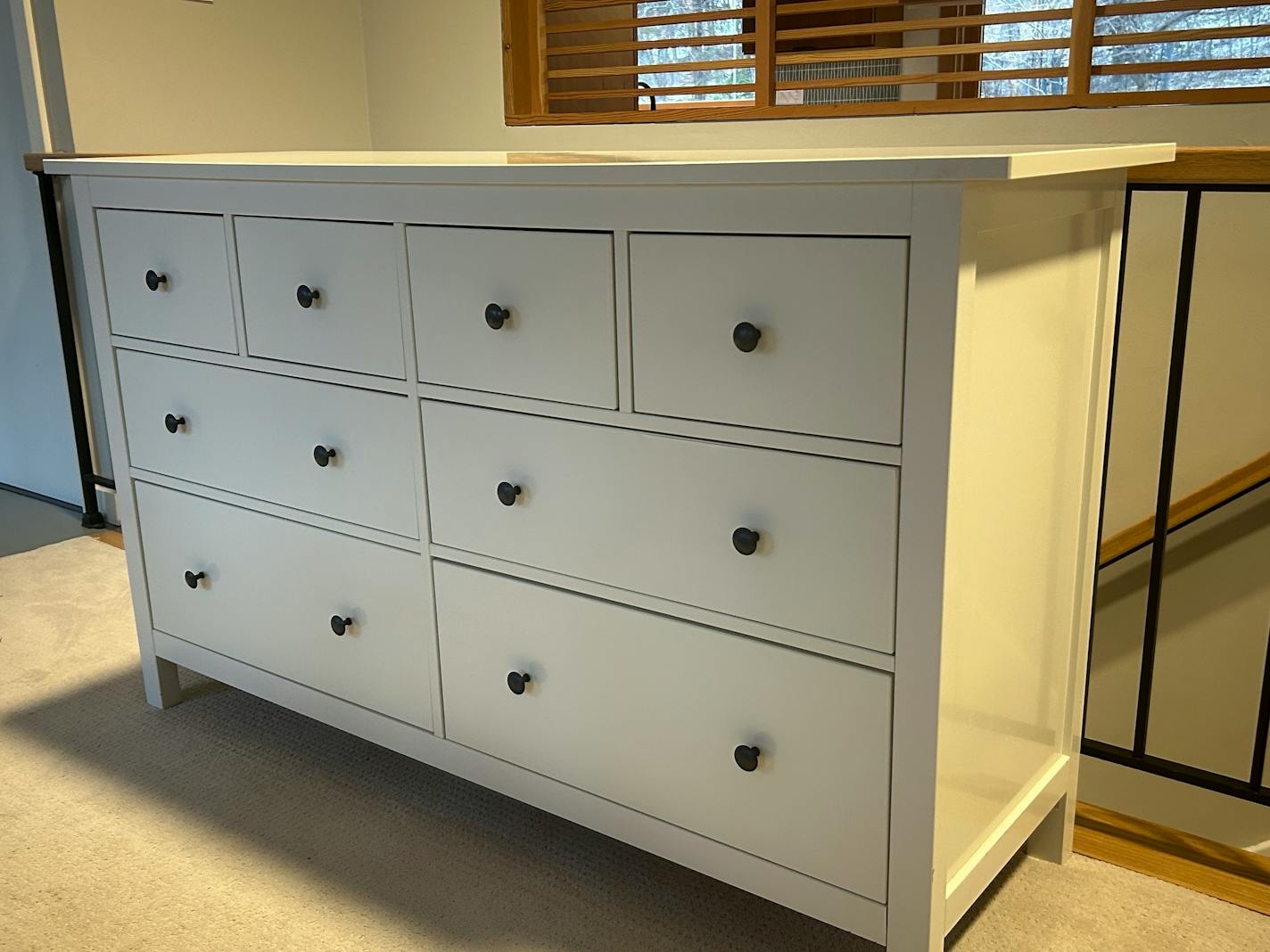 A large modern white dresser with eight drawers sits in a living area beside a staircase with metal railings.