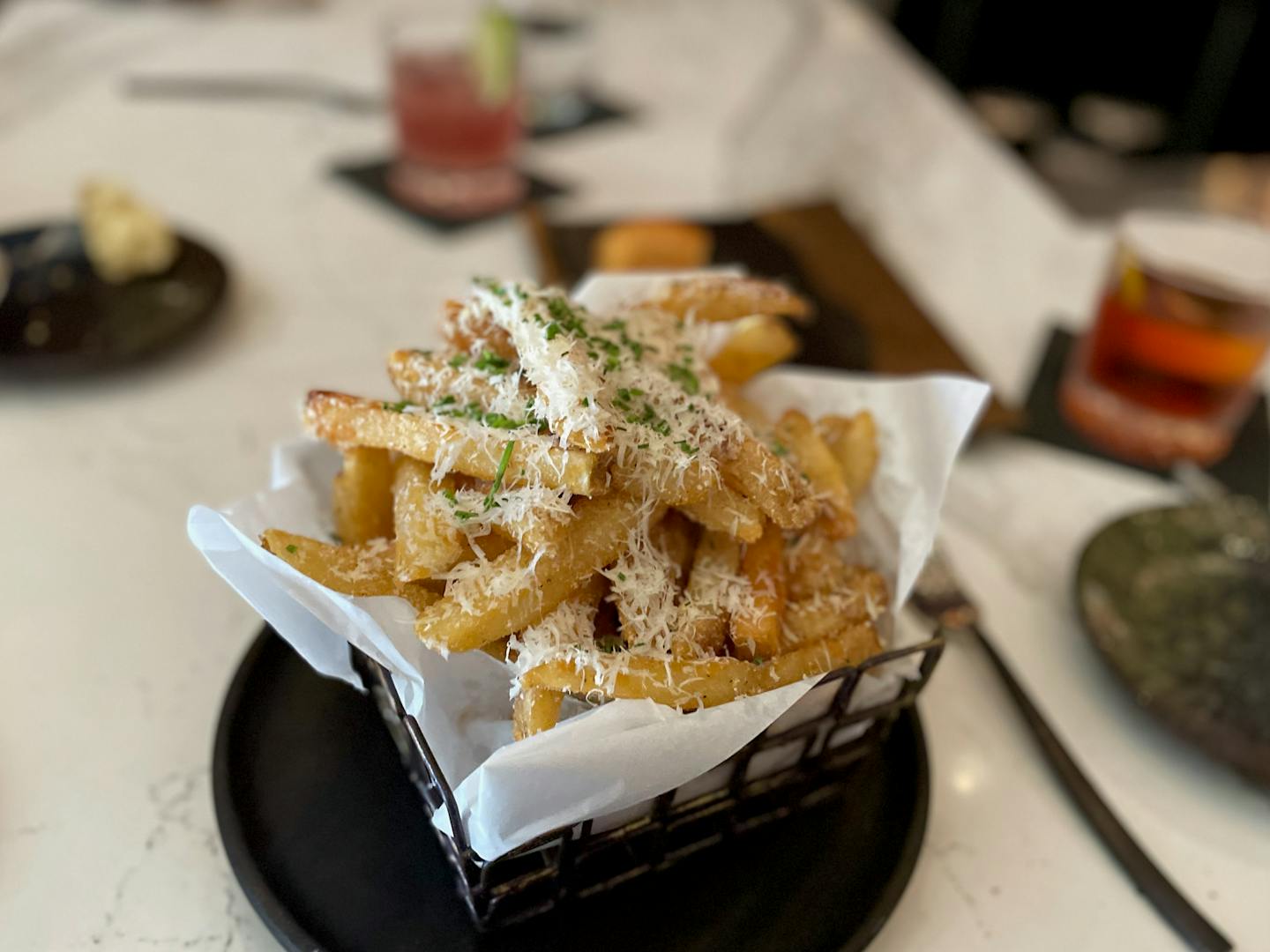 Boketto's French fries are served with a white napkin lined black metal basket and topped with parmesan cheese and truffle oil. Photographed on the bar with two cocktails in the background.