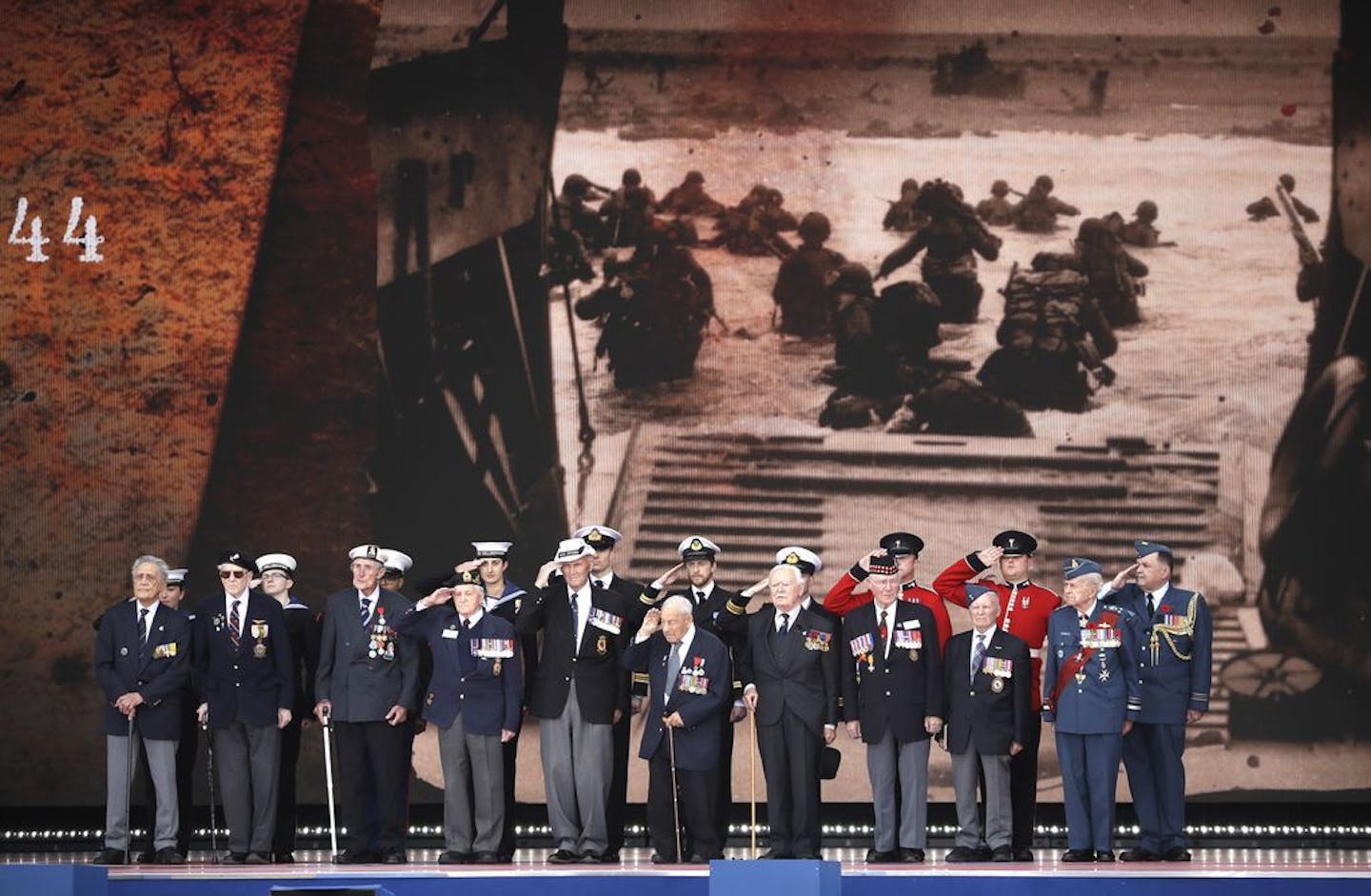 D-Day veterans are honored on stage at an event in England marking tomorrow's 75th anniversay of the Normandy invasion, depicted in the image behind them.