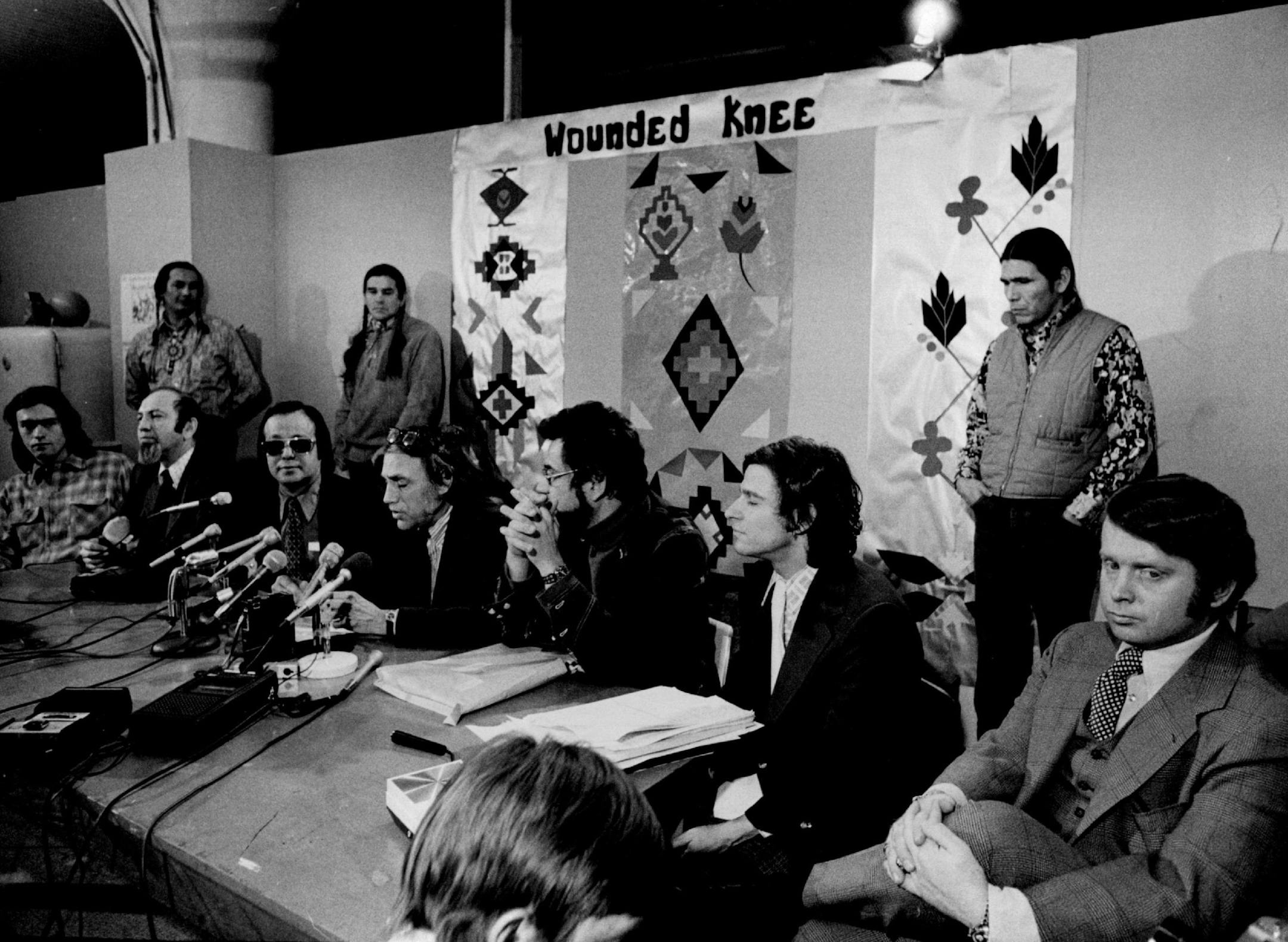 January 8, 1974: Defense attorneys for Russell Means, standing, left, and Dennis Banks, standing, right, outlined their initial court-room strategy Monday for the trial of the two American Indian Movement (AIM) leaders that begins today in St. Paul. The lawyers were, seated from left, Kenneth Tilsen, Ramon Roubideaux, William Kunstler, Mark Lane, Larry Leventhal and Robert Bryan. Standing between Means and Banks was AIM leader Clyde Bellecourt.