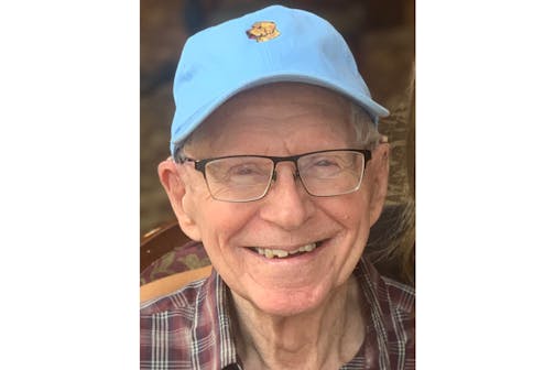 An elderly man wearing a blue baseball cap looks at the camera and smiles.