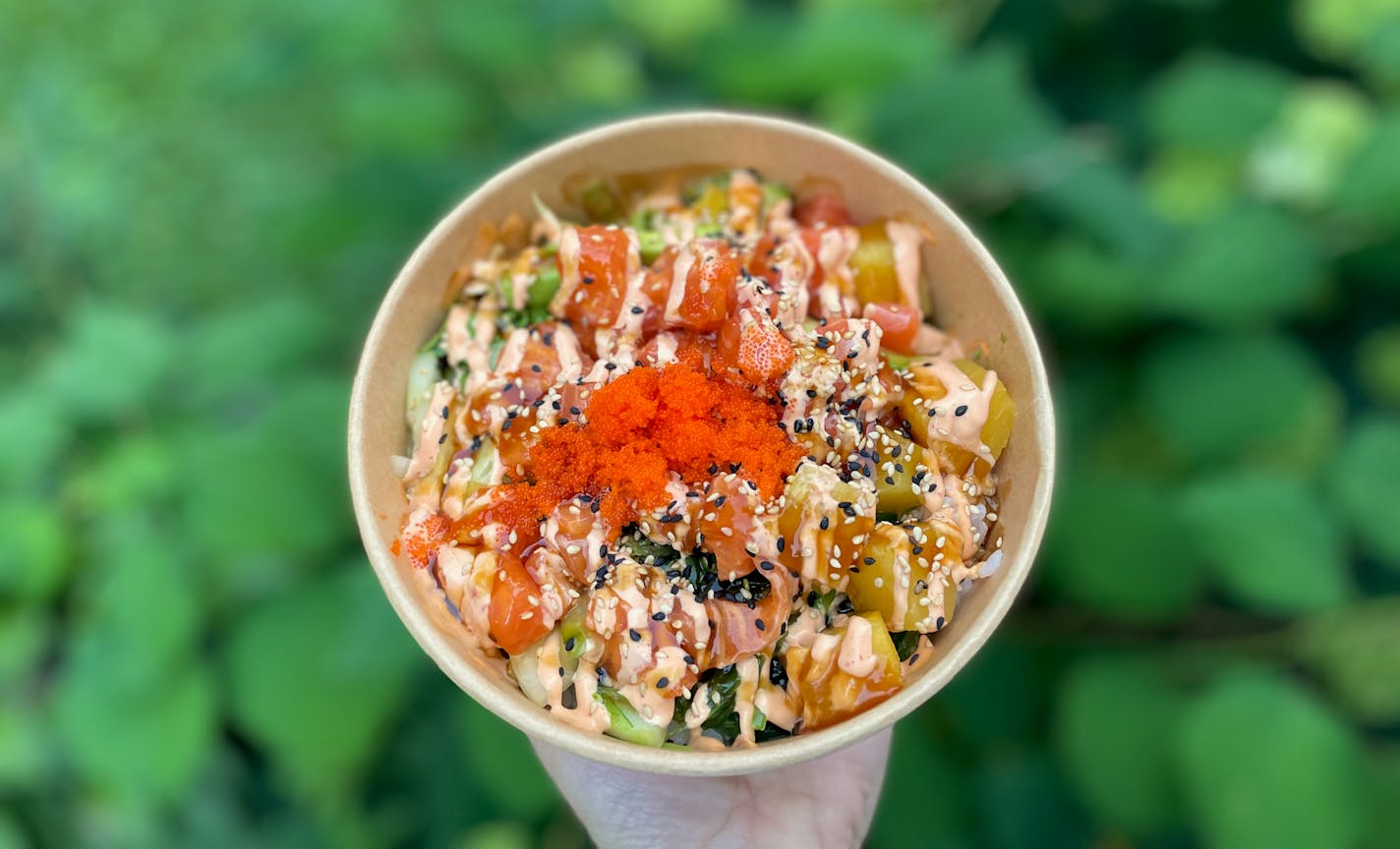 A paper bowl holds diced salmon, tuna, seaweed salad, pineapple and drizzled with spicy mayo. Garnished with bright orange trout roe. Held in front of an ivy covered background.