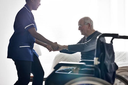 iStock Female caregiver helping senior man get up from bed at home.