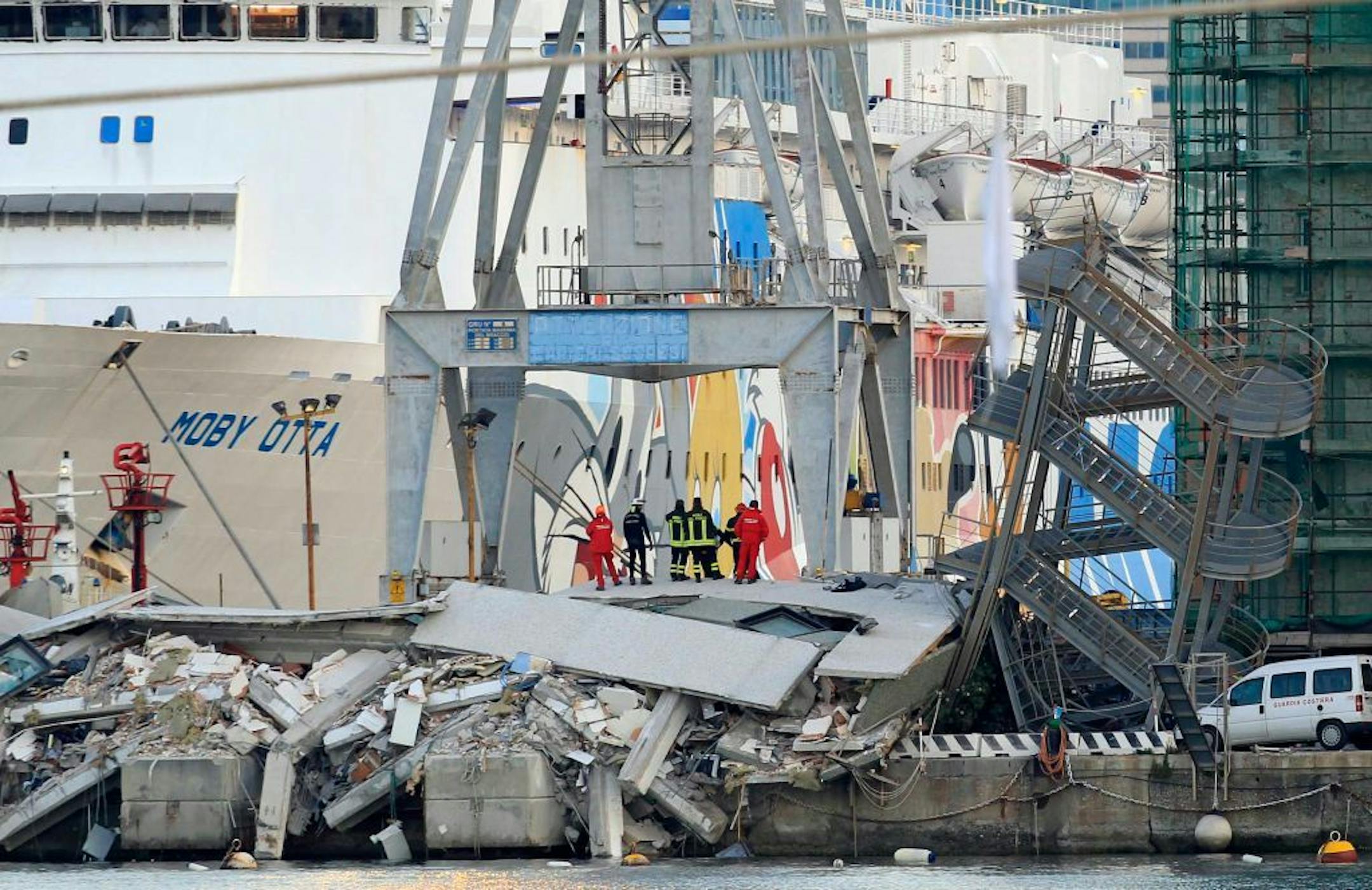 Rescue personnel stand on rubble next to a tilted staircase, part of a control tower which collapsed after a cargo ship slammed into it during a shift change Tuesday night in the port of Genoa, northern Italy, killing at least five people, Wednesday, May 8, 2013. By Wednesday morning, all that was left of the control tower was the mangled exterior staircase, tilted to its side. The tower itself, which was located on the very edge of a dock jutting out into the harbor, was either in the water or