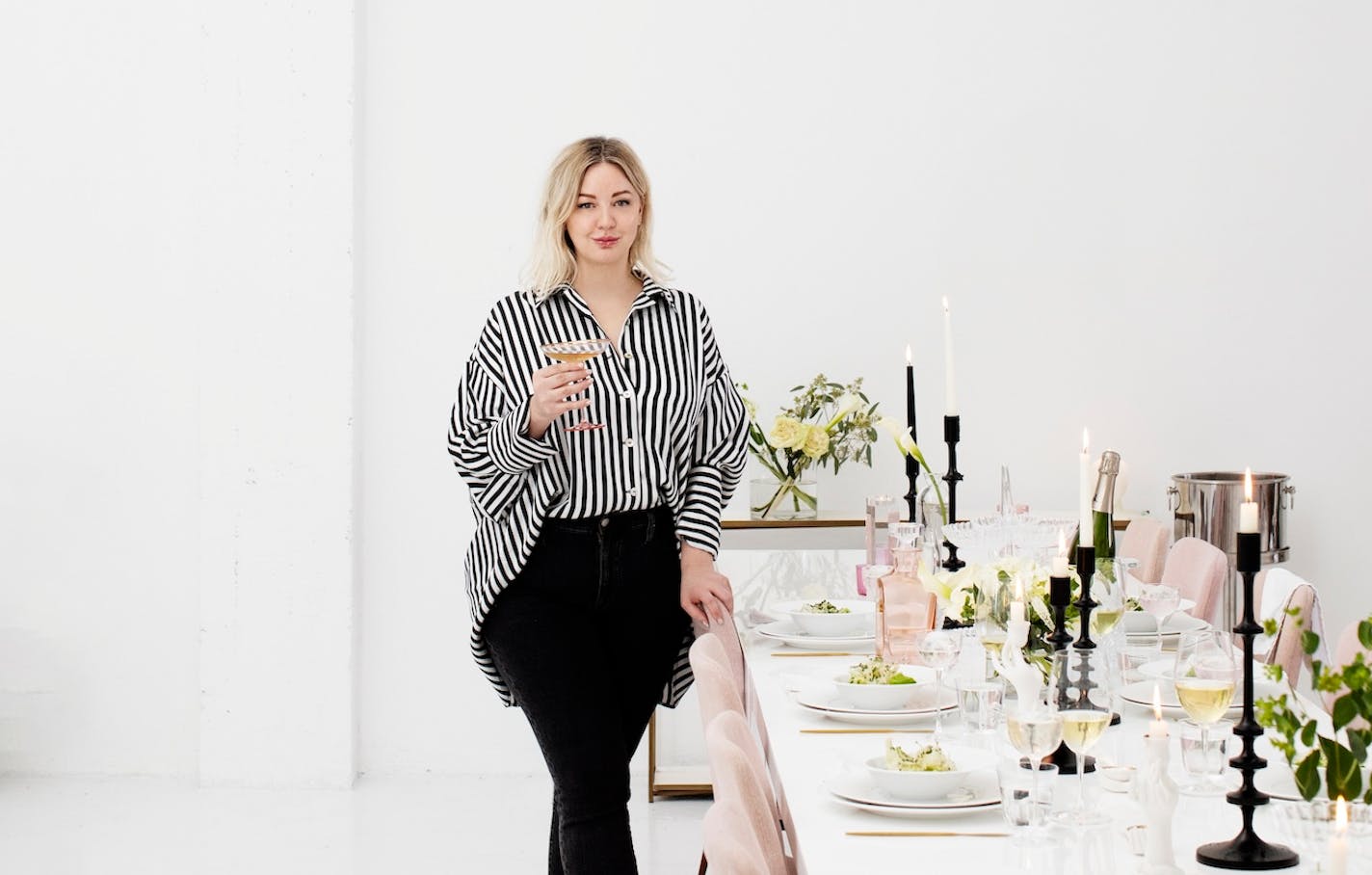 Jamie Malone stands at the head of a long, set table inside a white painted North Loop loft, holding a pink coup glass filled with champagne. She wears a black and white striped button up shirt, black jeans, and a slight, half-turned up smile.