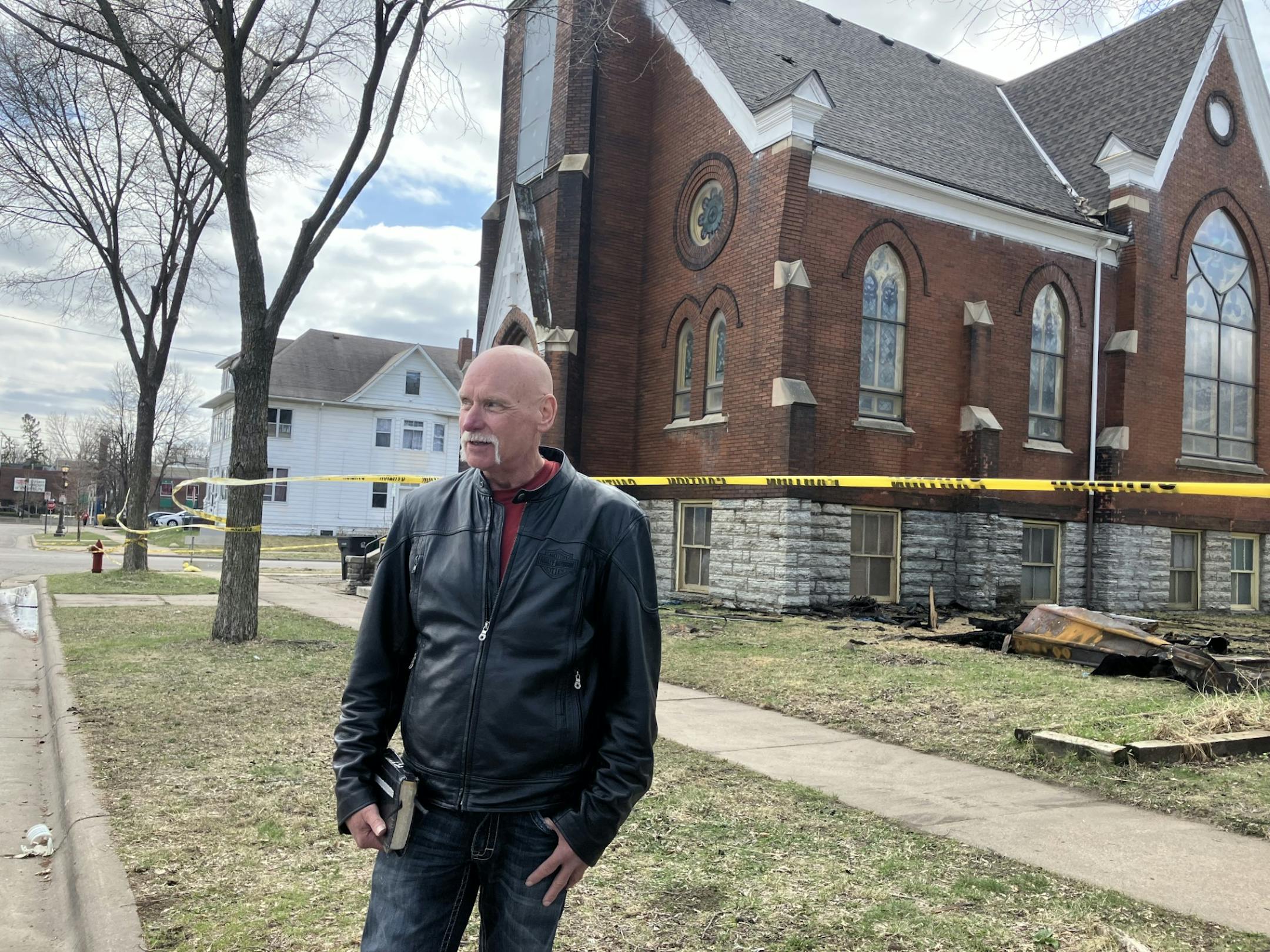 A man in a leather jacket stands in front of a brick church surrounded by yellow caution tape.