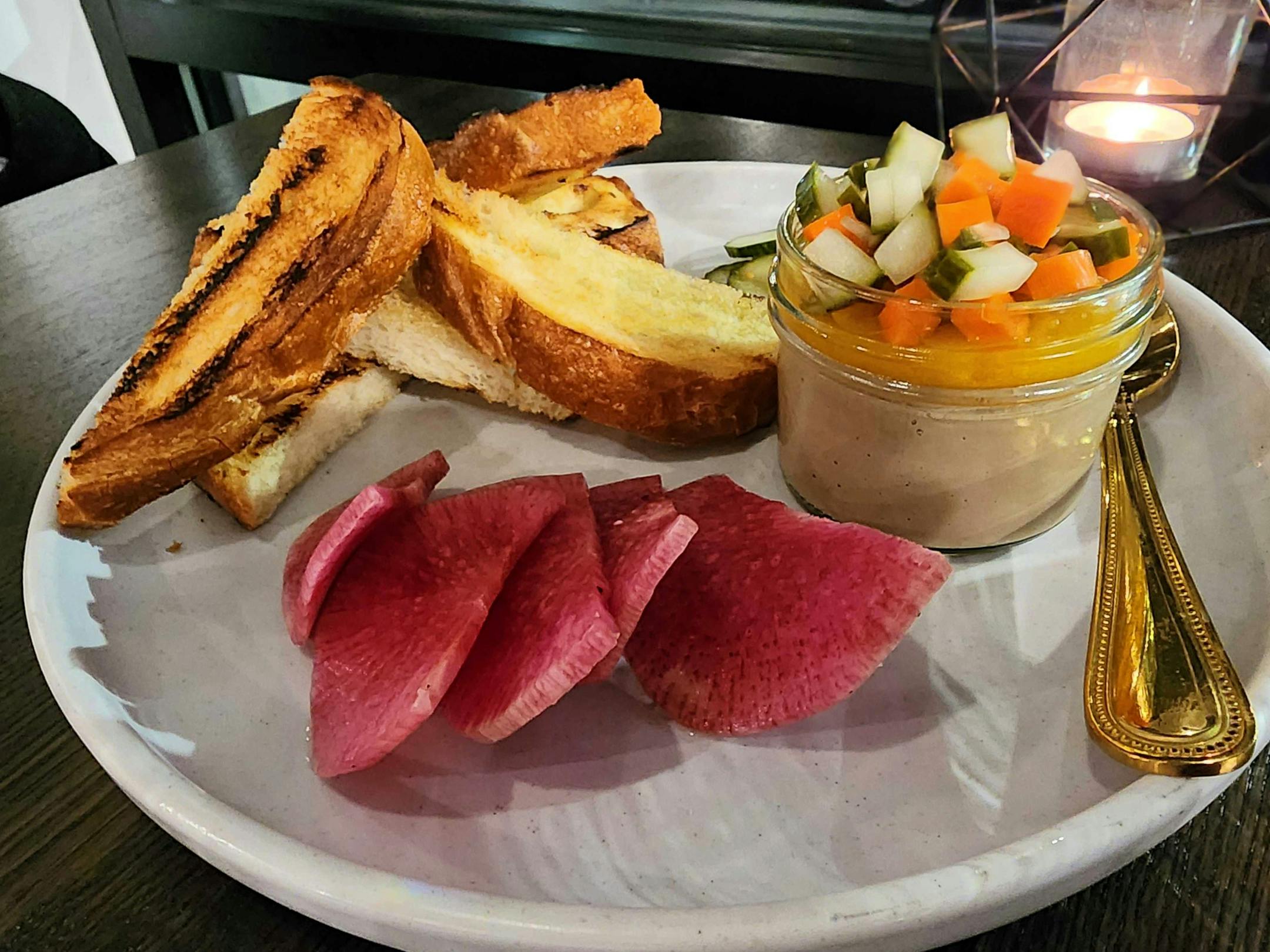A white plate with a jar filled with light brown chicken liver mousse topped with diced cucumbers and carrots, and toast points