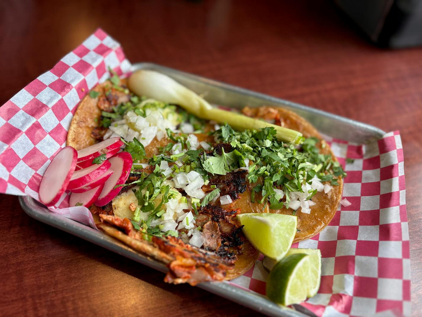 A metal tray with three tacos of crispy al pastor topped with onions, cilantro, limes, radish and a grilled green onion.