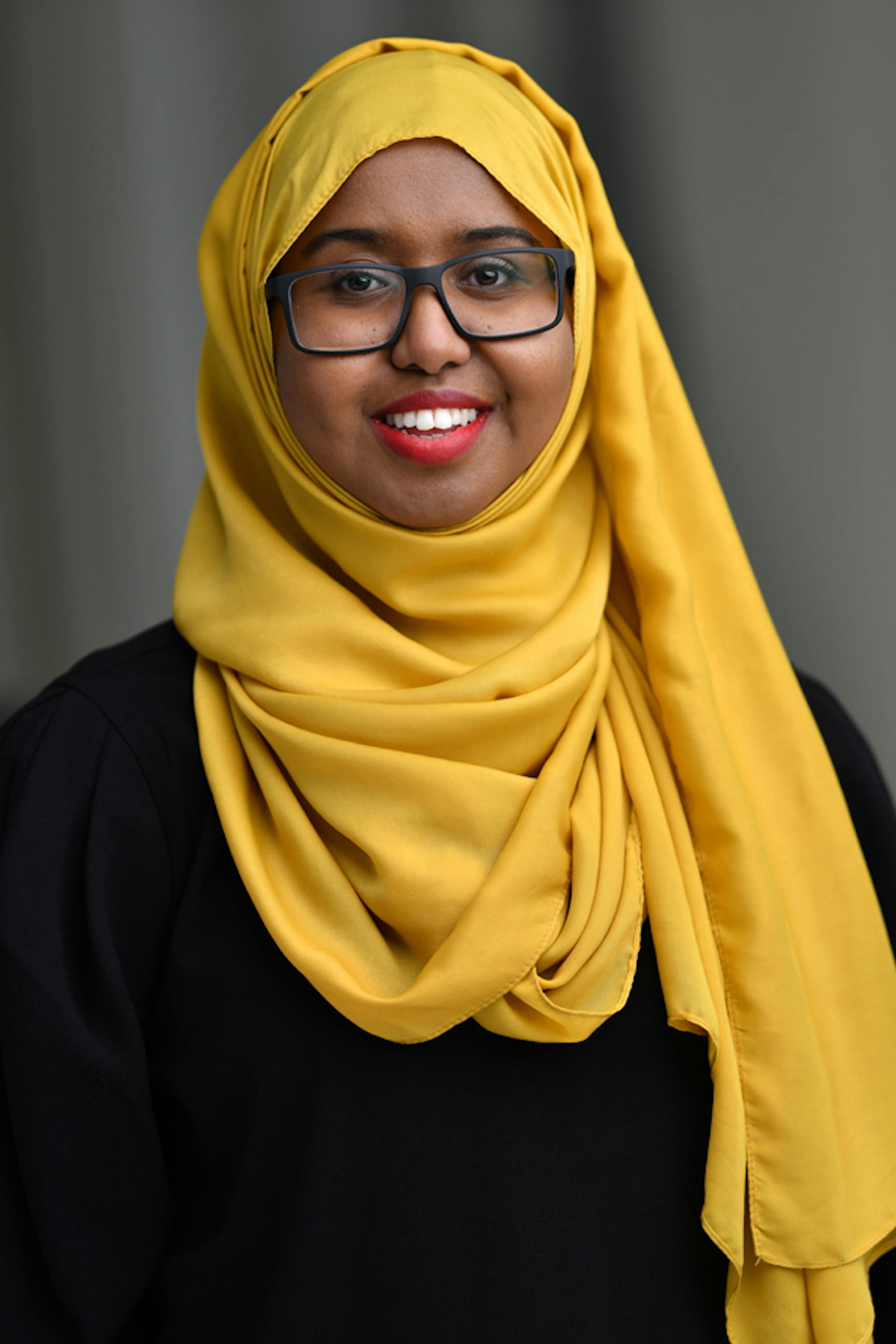A Somali American woman wearing a bright mustard-color hijab, dark framed glasses and pink lipstick smiles for the camera.