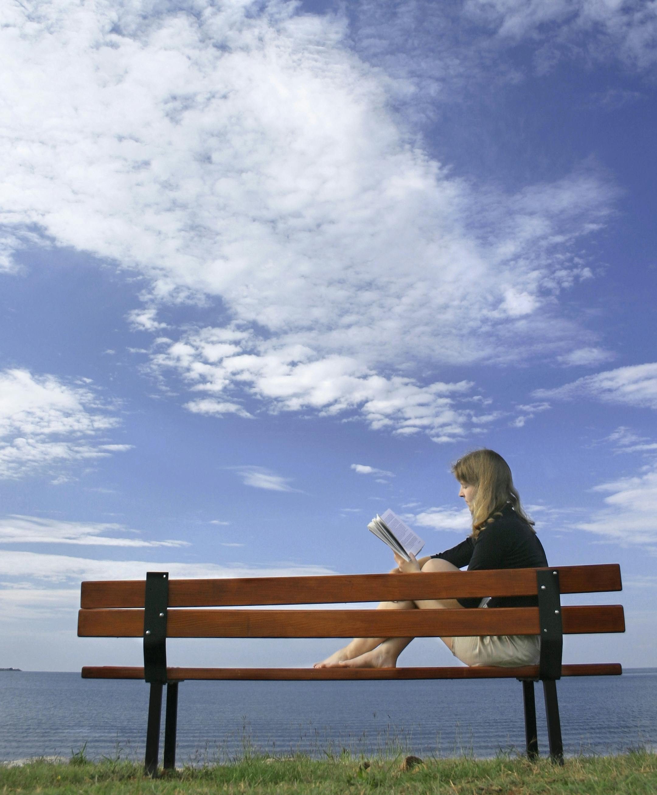istock Woman on bench, lake, reading book, blue sky