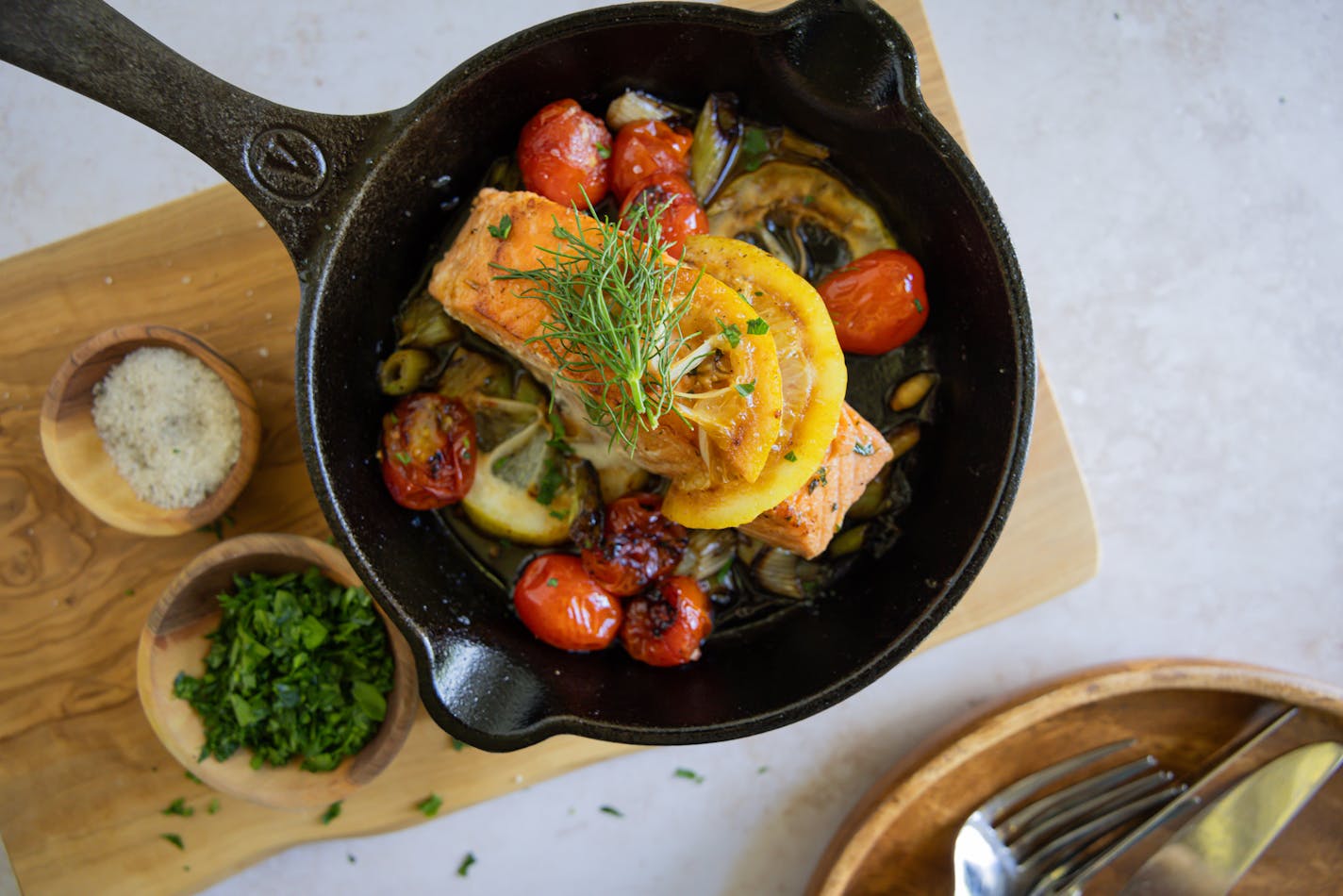 A cast-iron skillet with a salmon fillet on a bed of roasted cherry tomatoes, herbs and lemon and topped with additional herbs and lemon slices.